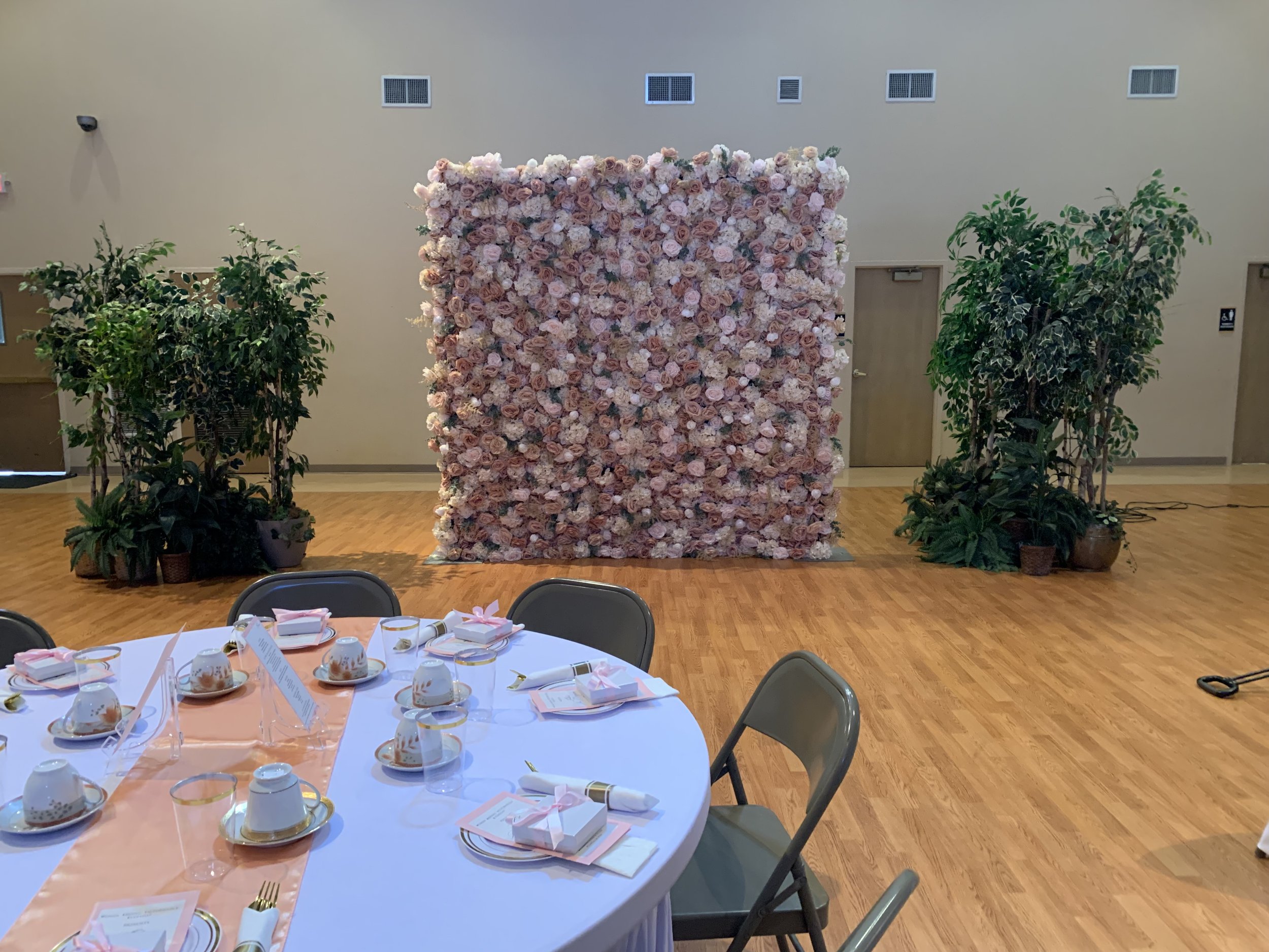 Decorated event space with a square wall of pink and white roses, two potted plants on either side, and a round table set for a celebration in the foreground.