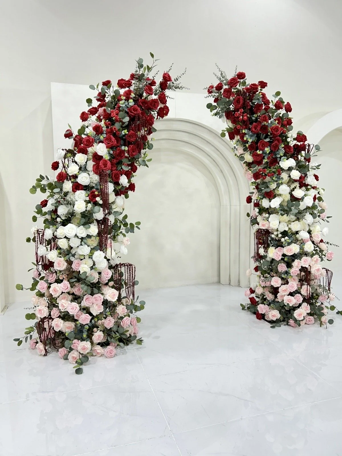 A floral arch decoration with red, white, and pink roses, greenery, and hanging bead accents on a white background.