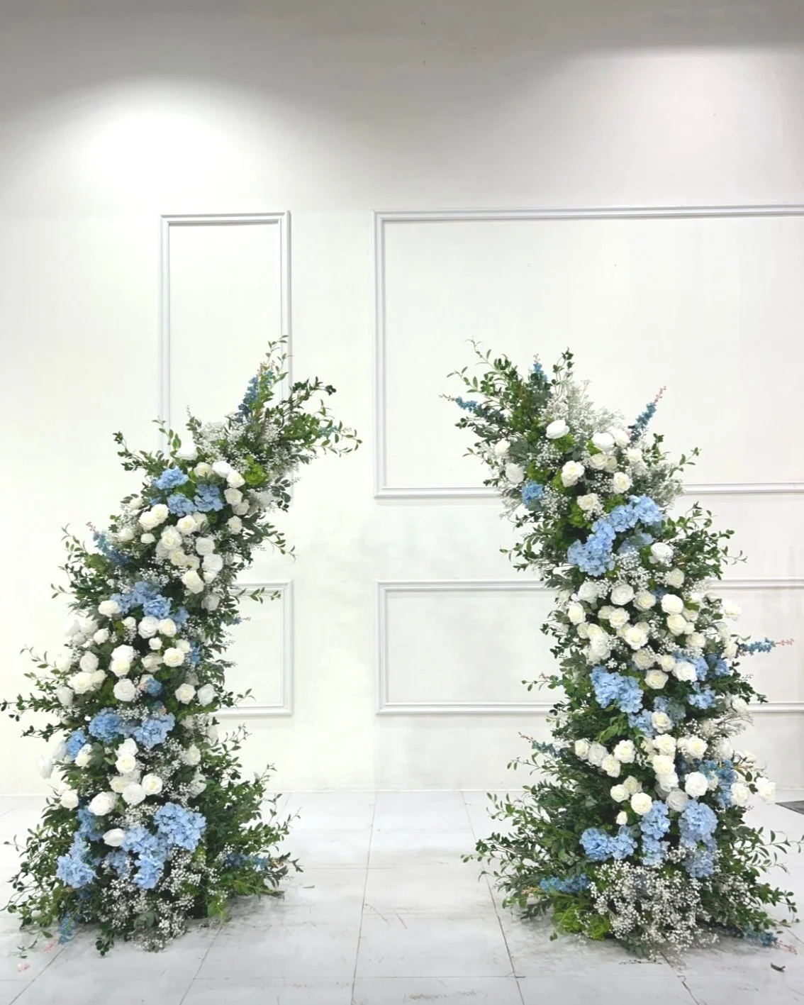 Two large floral arrangements with white and blue flowers and green foliage, arranged on the floor against a white wall with decorative paneling.