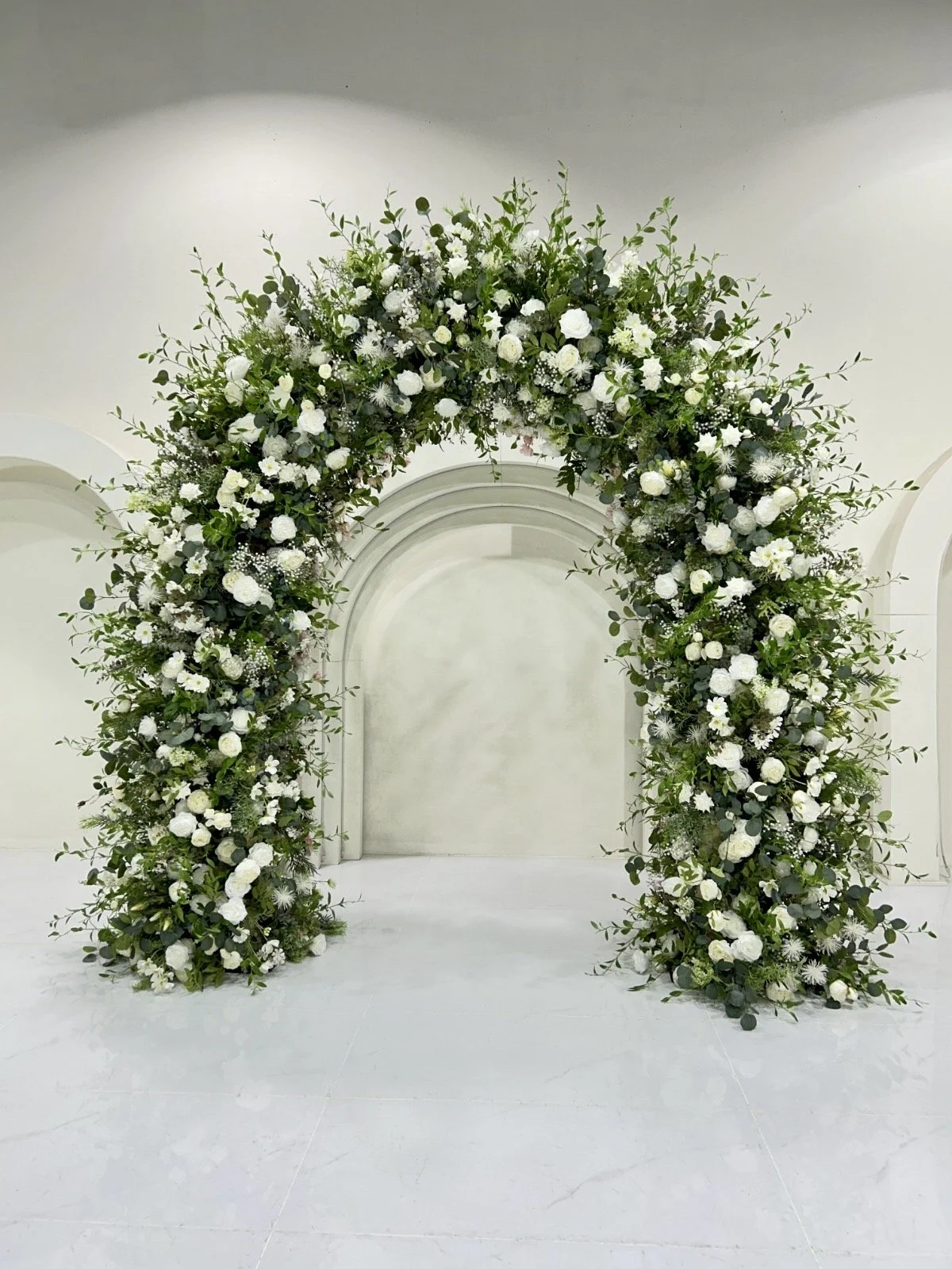 Large white floral arch with green foliage in an indoor setting.