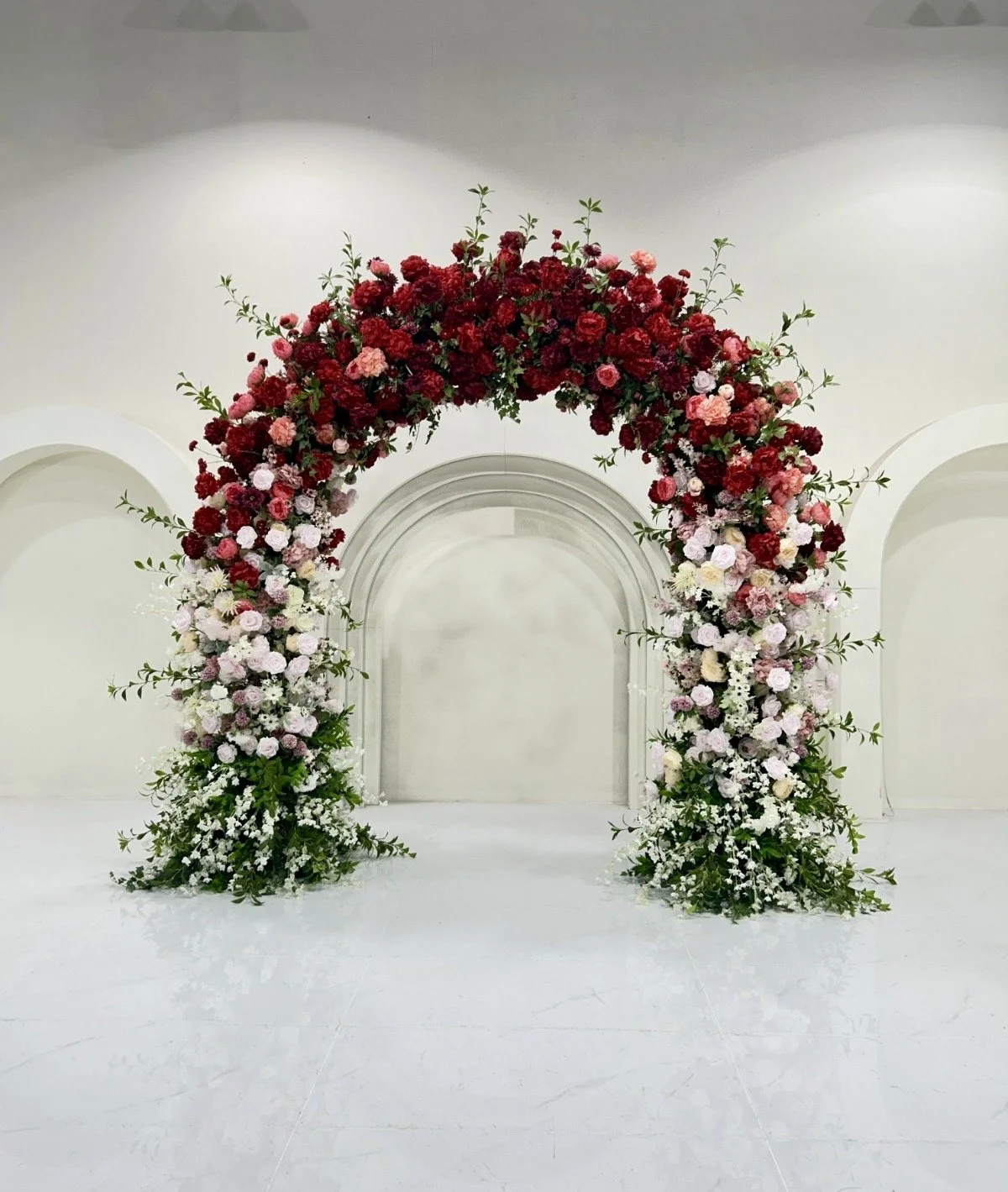 A floral wedding arch decorated with red, pink, and white flowers in a white indoor space.