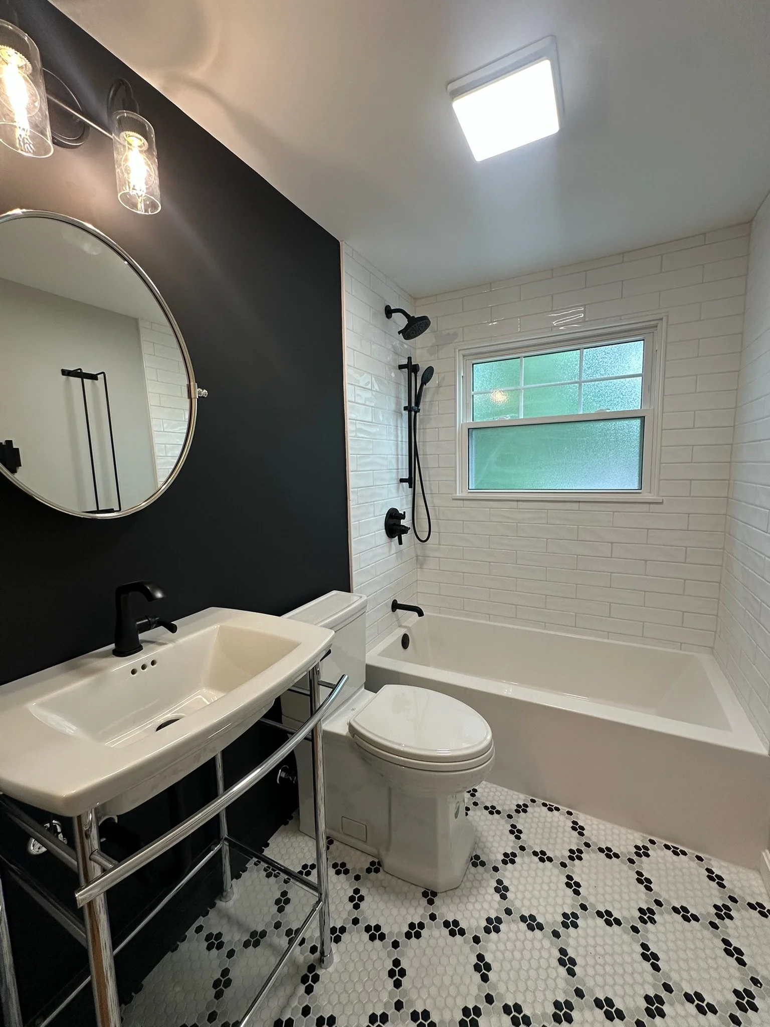 Modern bathroom with white subway tile walls, a black accent wall, a round mirror, a small white sink, a toilet, a bathtub with a black showerhead, and a large window with frosted glass. The floor has white hexagonal tiles with black accents.