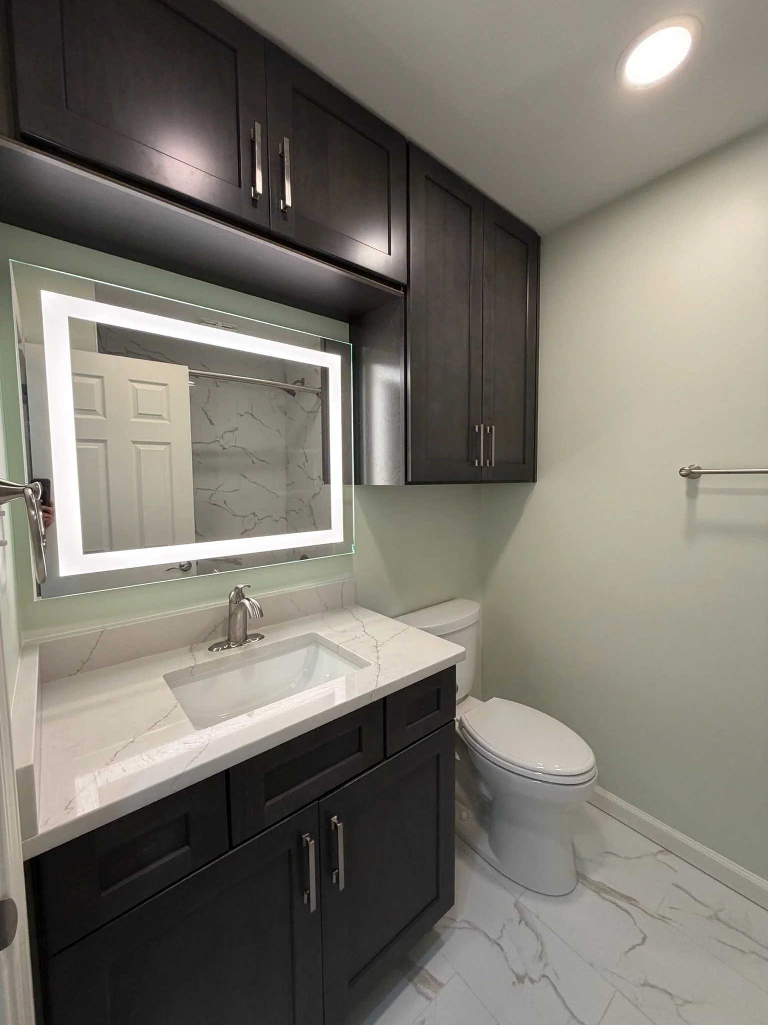 Modern bathroom with dark wood cabinets, marble countertop with sink, illuminated mirror, toilet, light green walls, and marble tile flooring.