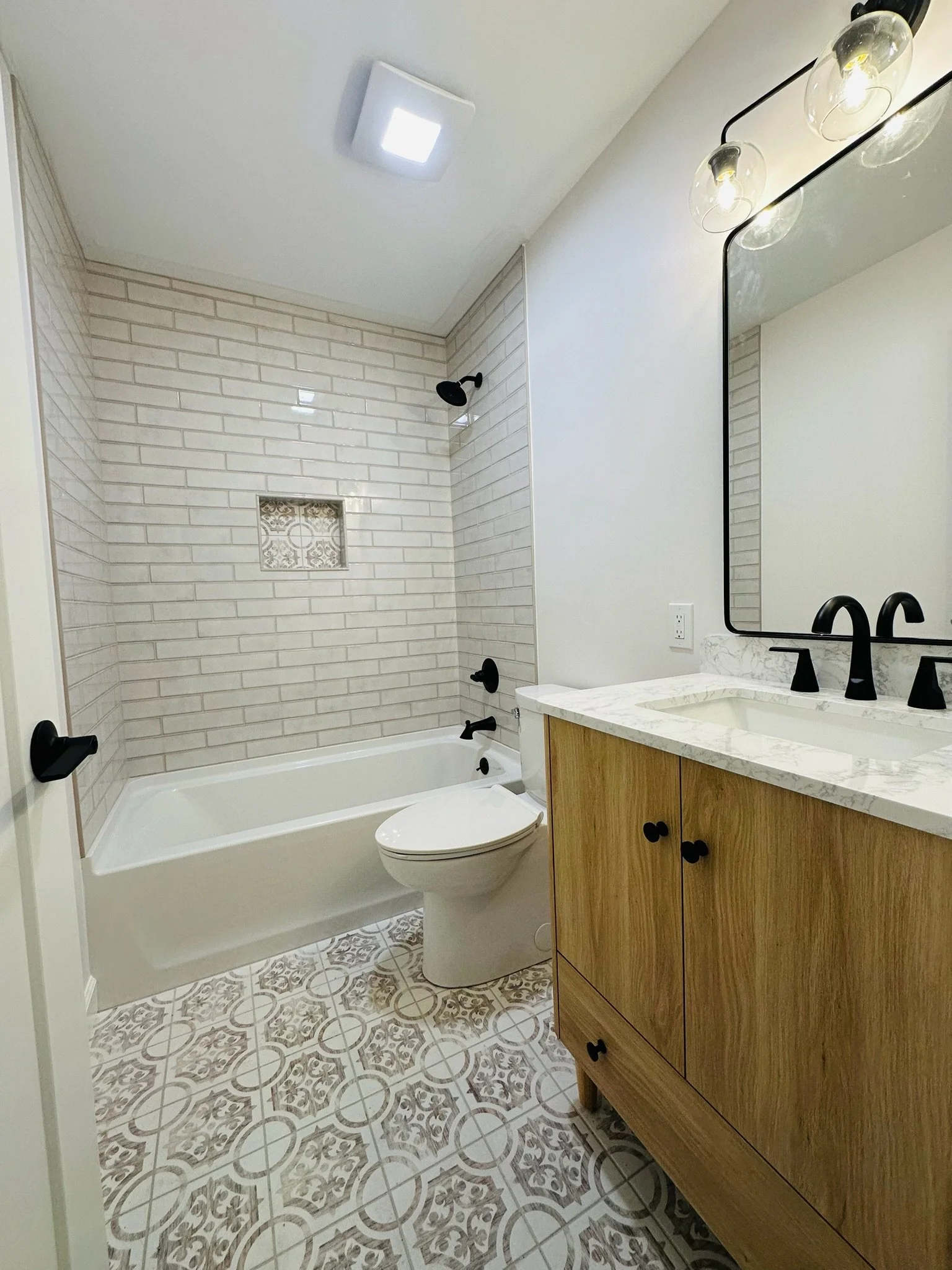 Bathroom with a bathtub, white brick tile walls, a toilet, a wooden vanity with marble countertop, black fixtures, and patterned floor tiles.