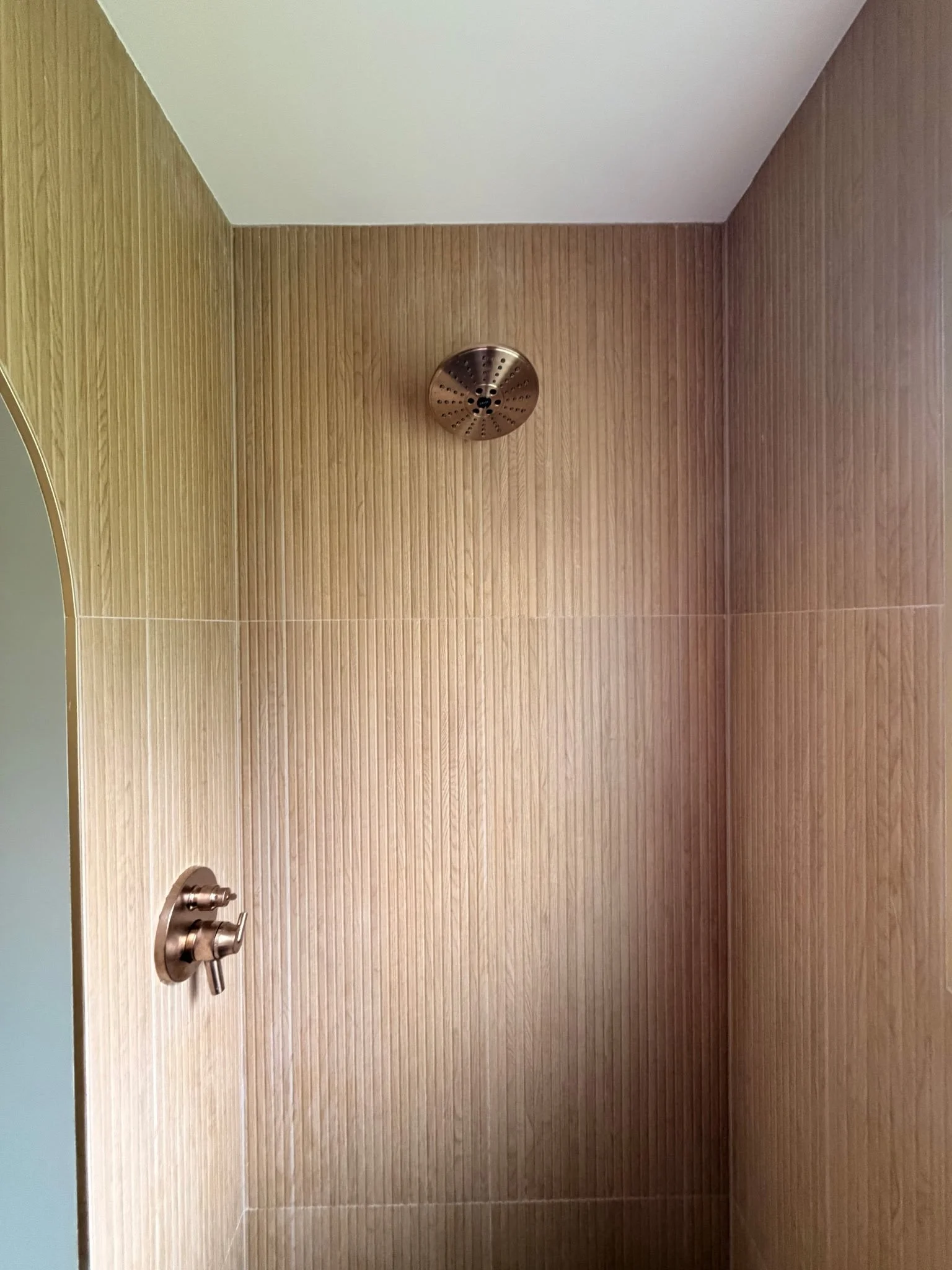 A shower with wood-textured wall tiles, a round metal showerhead, and a bronze-colored shower handle.