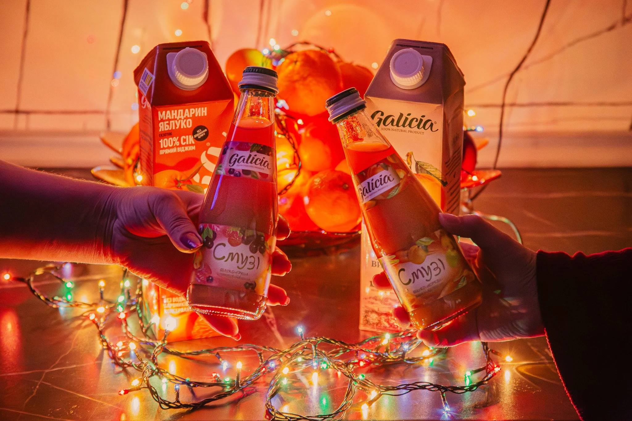 Two people are holding bottles of fruit juice with a background of pumpkins and colorful string lights, creating a festive autumn or Thanksgiving scene.