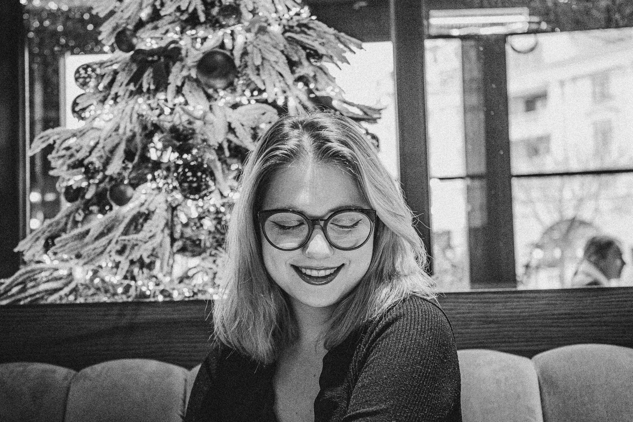 A woman with glasses and shoulder-length hair smiling with her eyes closed, sitting indoors with a Christmas tree in the background, decorated with ornaments.