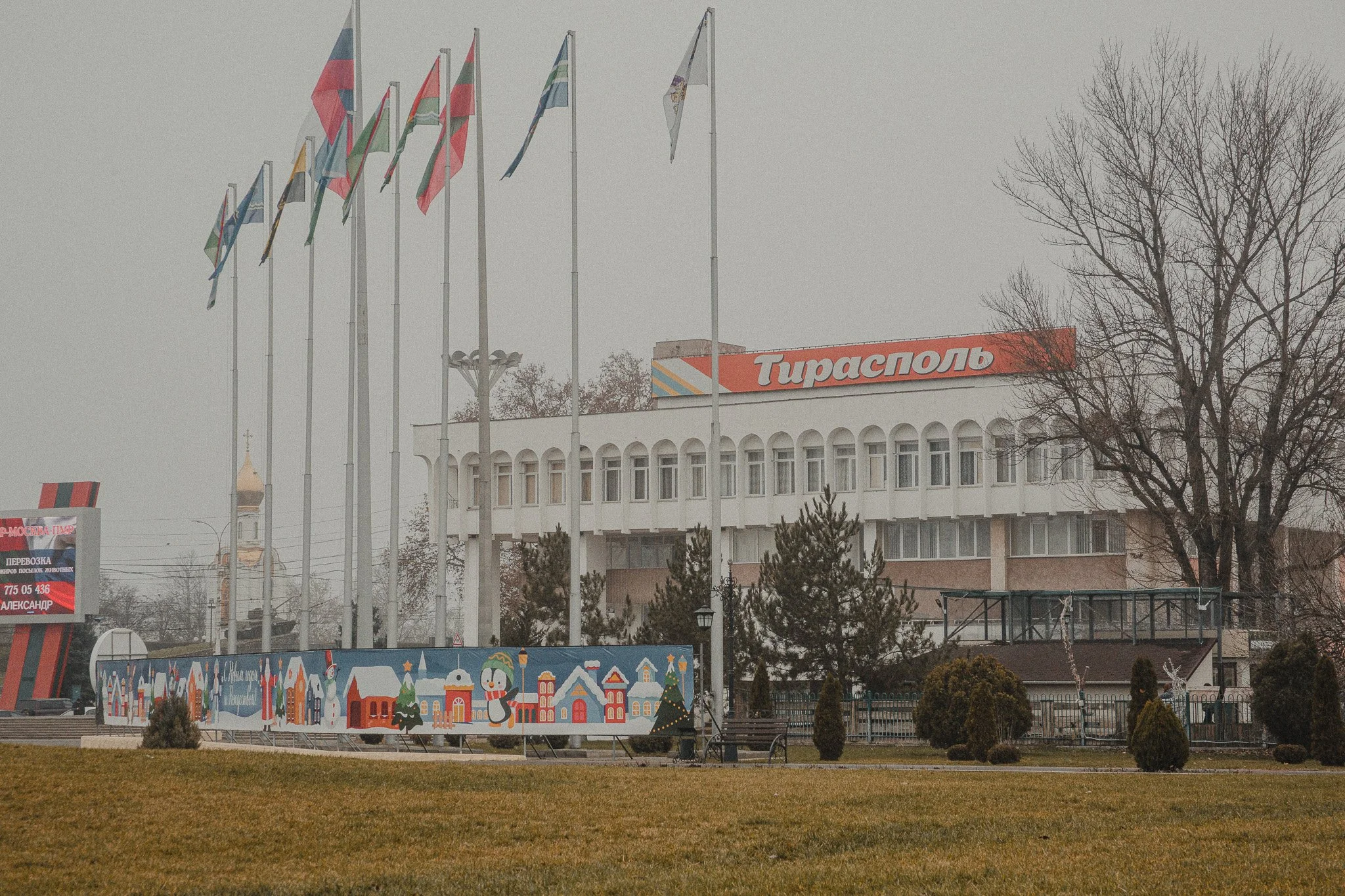 A building with a large orange sign that reads 'Тирасполь' in Cyrillic, surrounded by several flagpoles with flags, some of which are blue, white, and red. There are trees, a colorful outdoor display with holiday-themed images, and a grass lawn in fr
