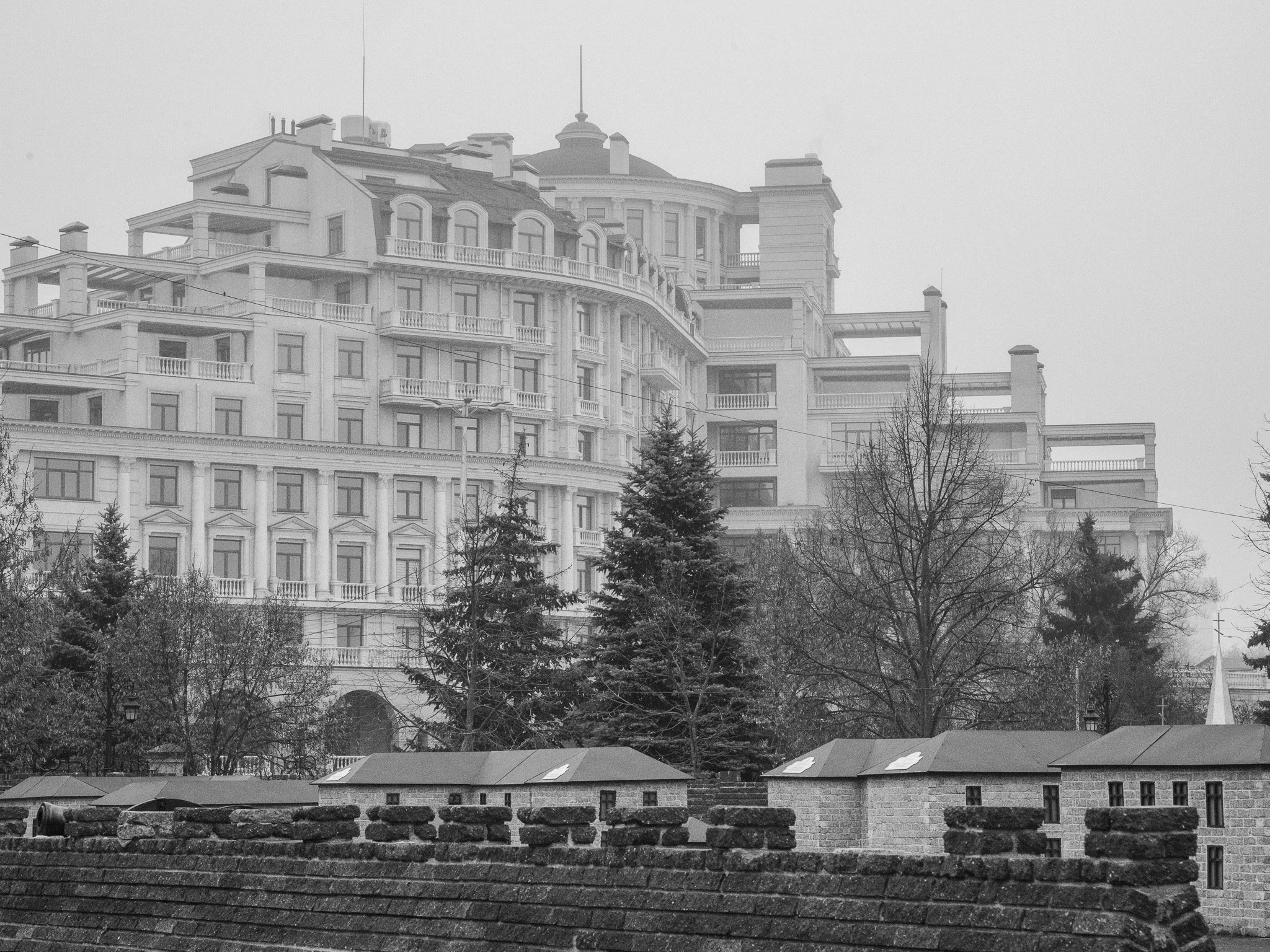 A large, ornate multi-story building with balconies and decorative architectural details, partially obscured by a layer of fog, with trees and a brick wall in the foreground.