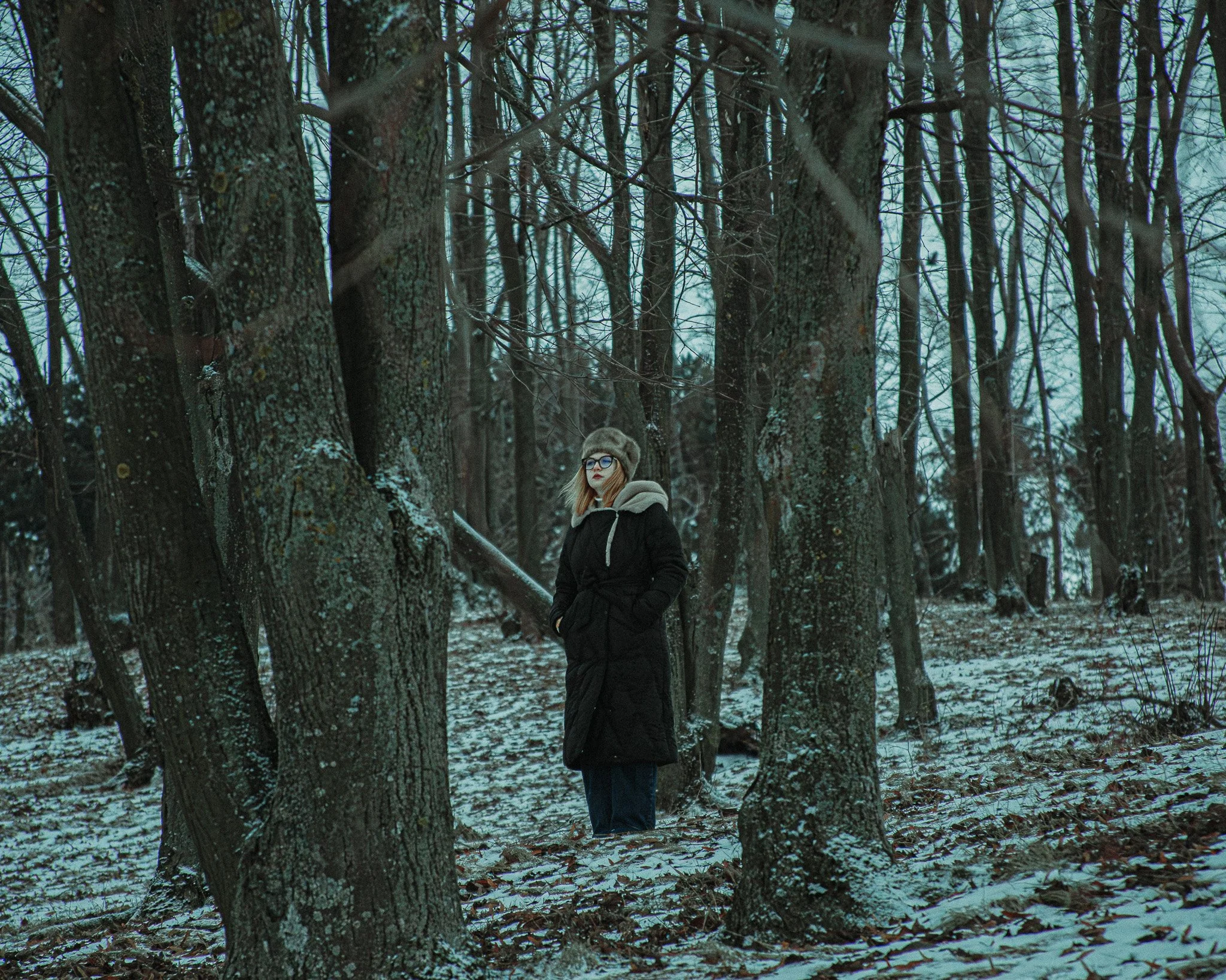 A woman standing alone in a snowy forest with tall, leafless trees, wearing a long black winter coat, a fur hat, glasses, and looking to the side.