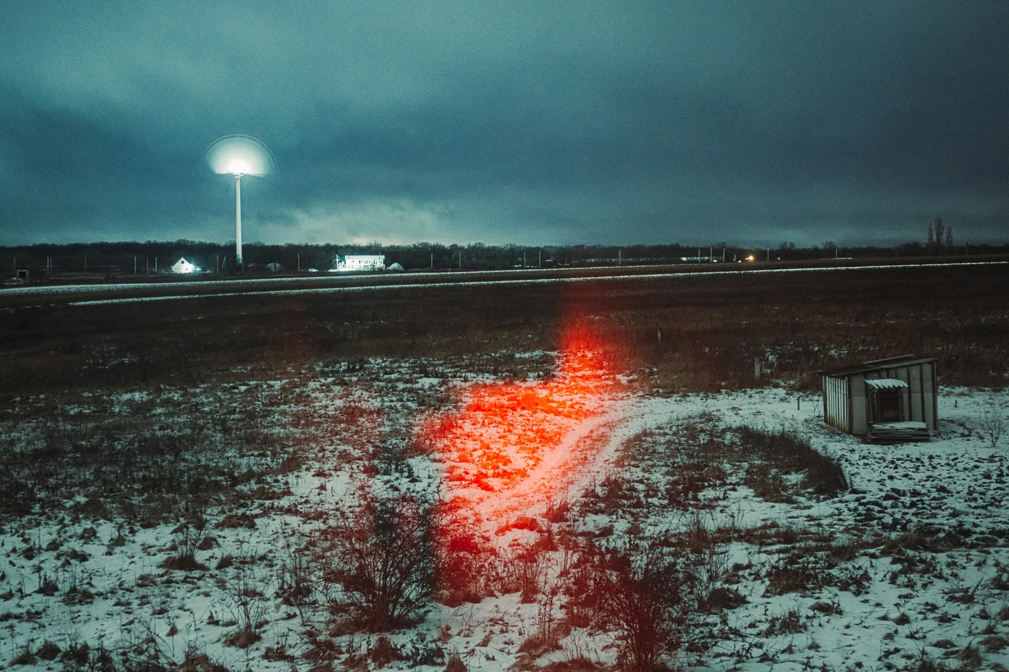 Nighttime rural scene with snow-covered ground, a small shed on the right, a red light on the ground, and a distant wind turbine with a white light. Dark sky with some clouds and a few illuminated buildings in the background.