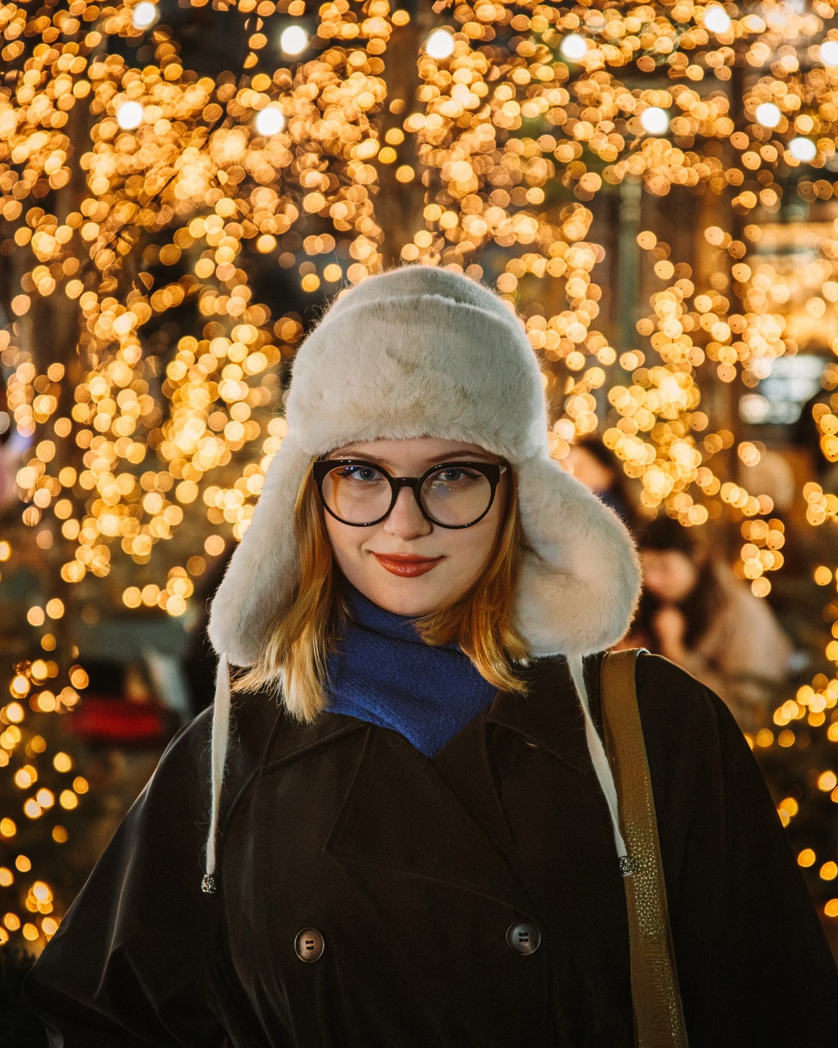 Young woman with glasses and red hair wearing a white furry hat and dark coat, standing in front of Christmas lights.