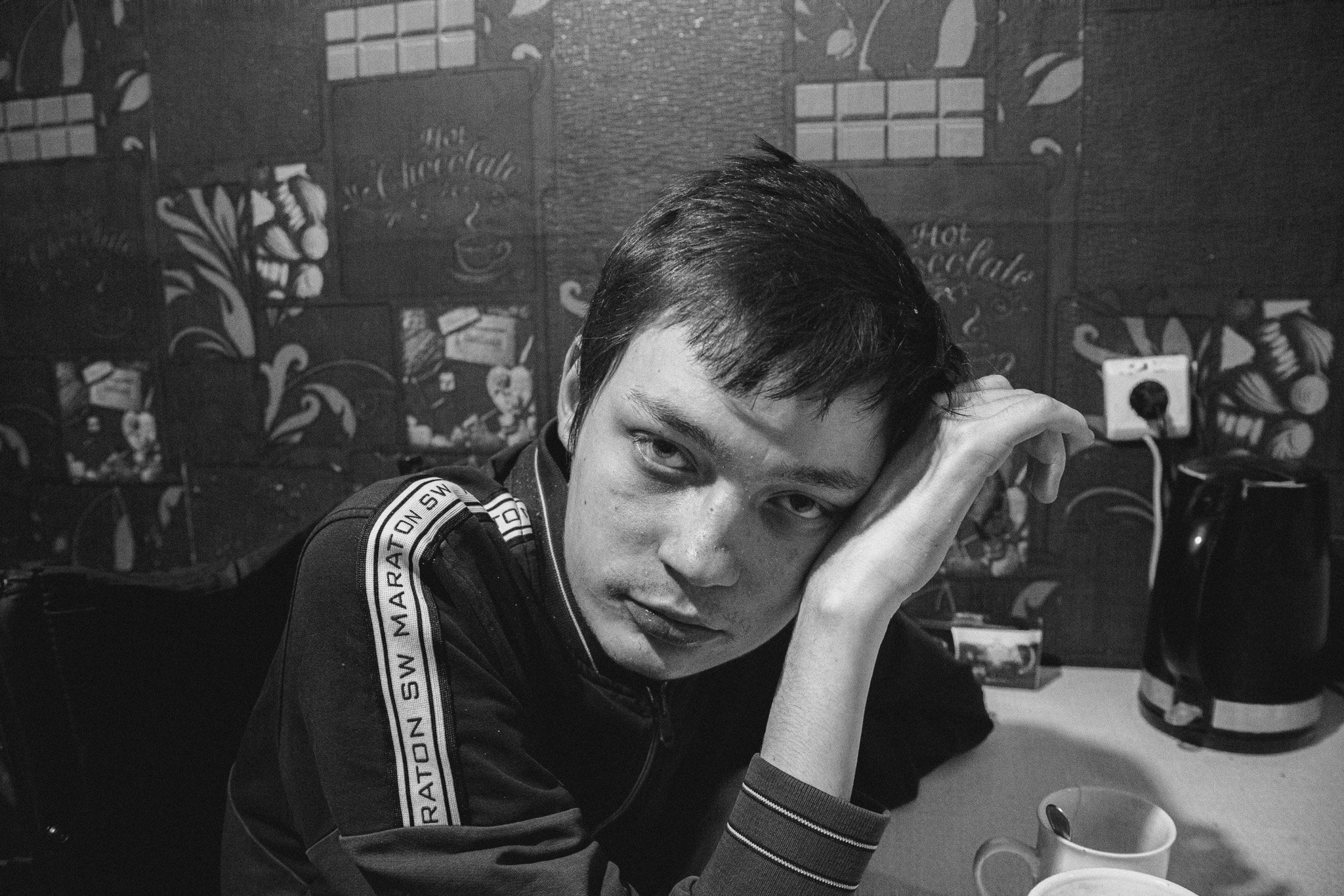 A young man with short dark hair and freckles resting his head on his hand at a table, looking at the camera. The background features a wall with a patterned mural or wallpaper. There is a coffee maker, a mug, and a power outlet visible on the table.
