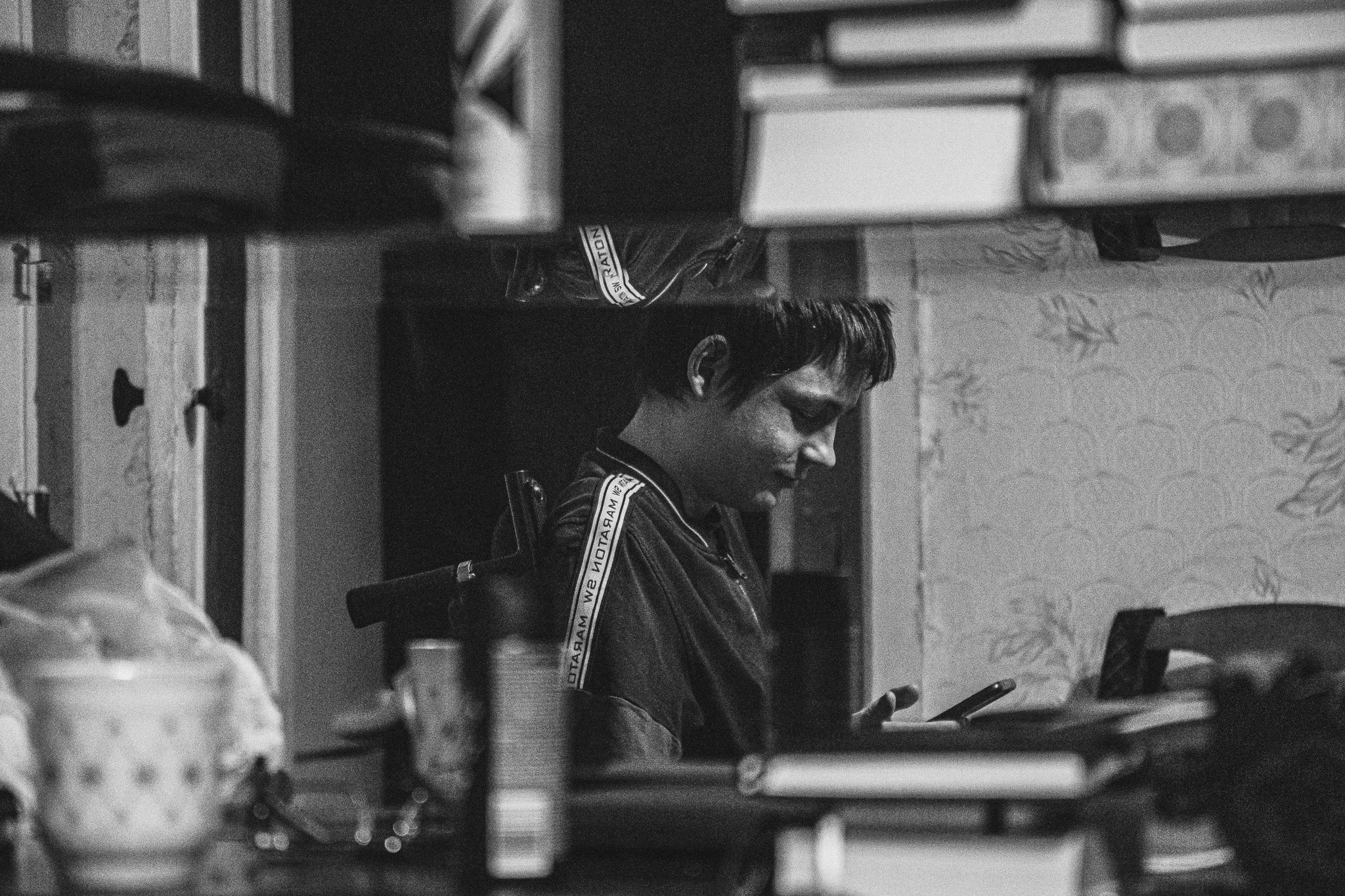 A young man sitting at a table, looking at his phone, in a room with shelves and containers in the background.