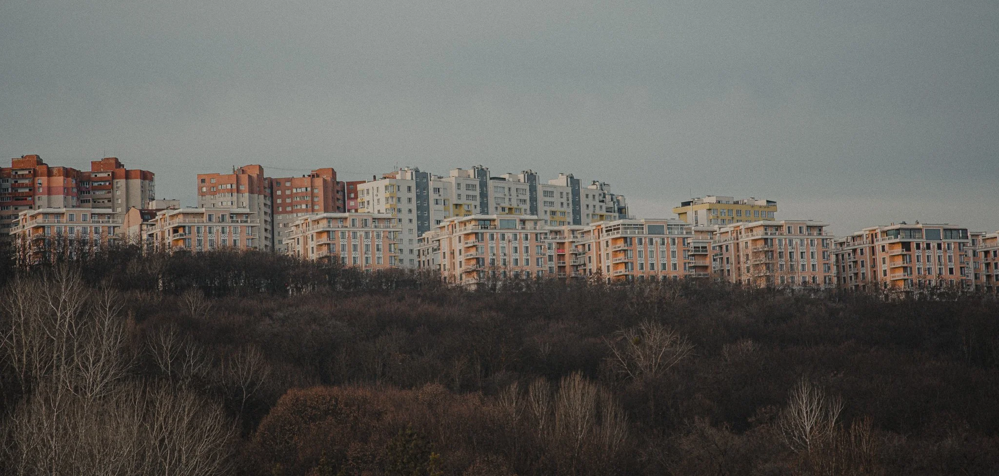 Multiple apartment buildings on a hillside during the daytime under an overcast sky, with leafless trees in the foreground.