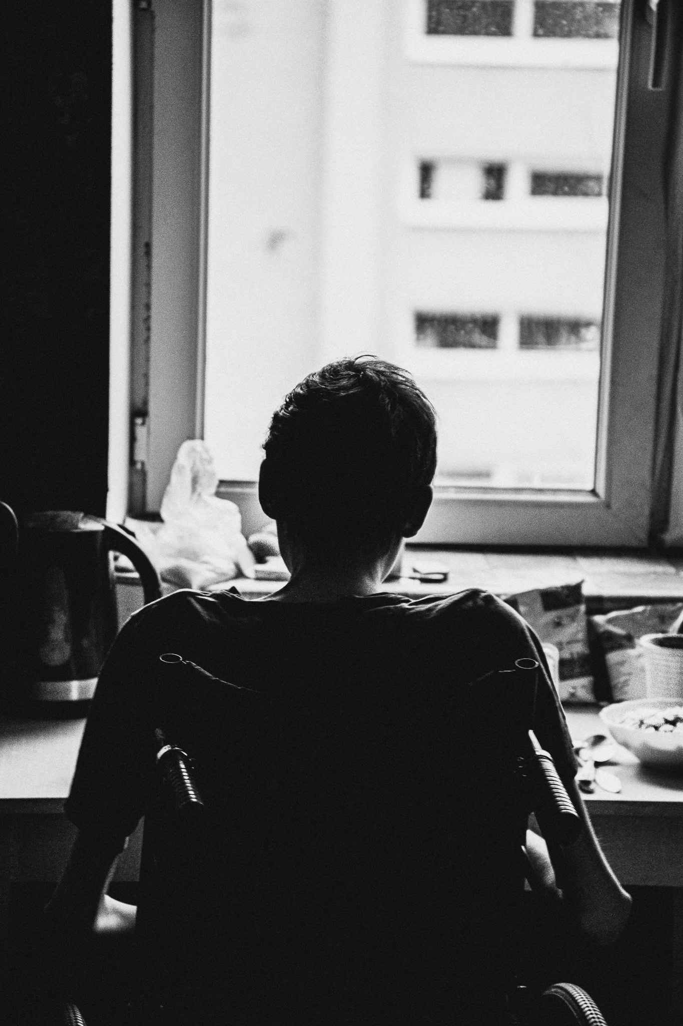 Silhouette of a person sitting in a wheelchair, facing a window with a building outside, in black and white.