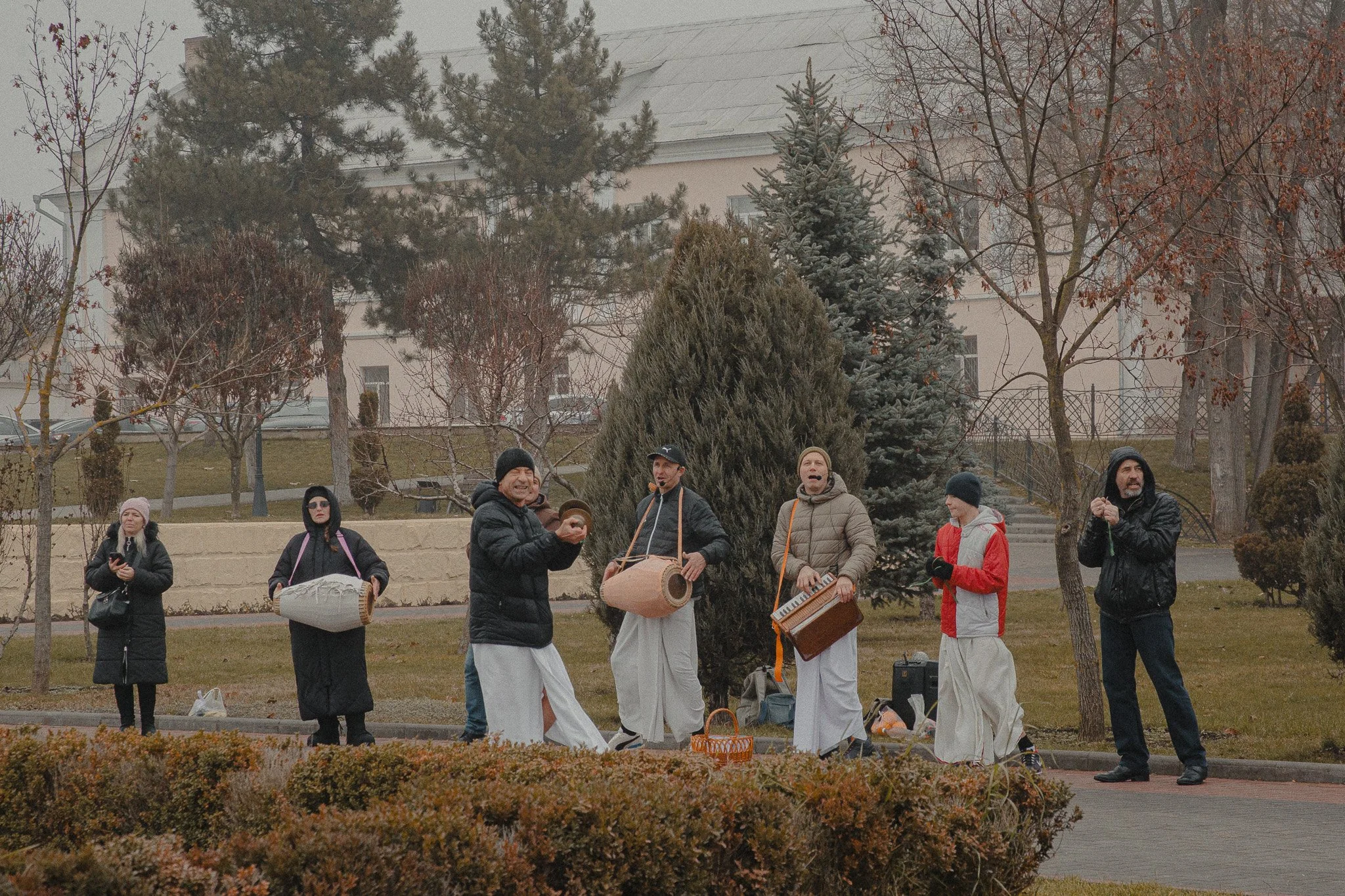 A group of people outdoors, some dressed in casual winter clothing, gather around musicians playing traditional instruments.