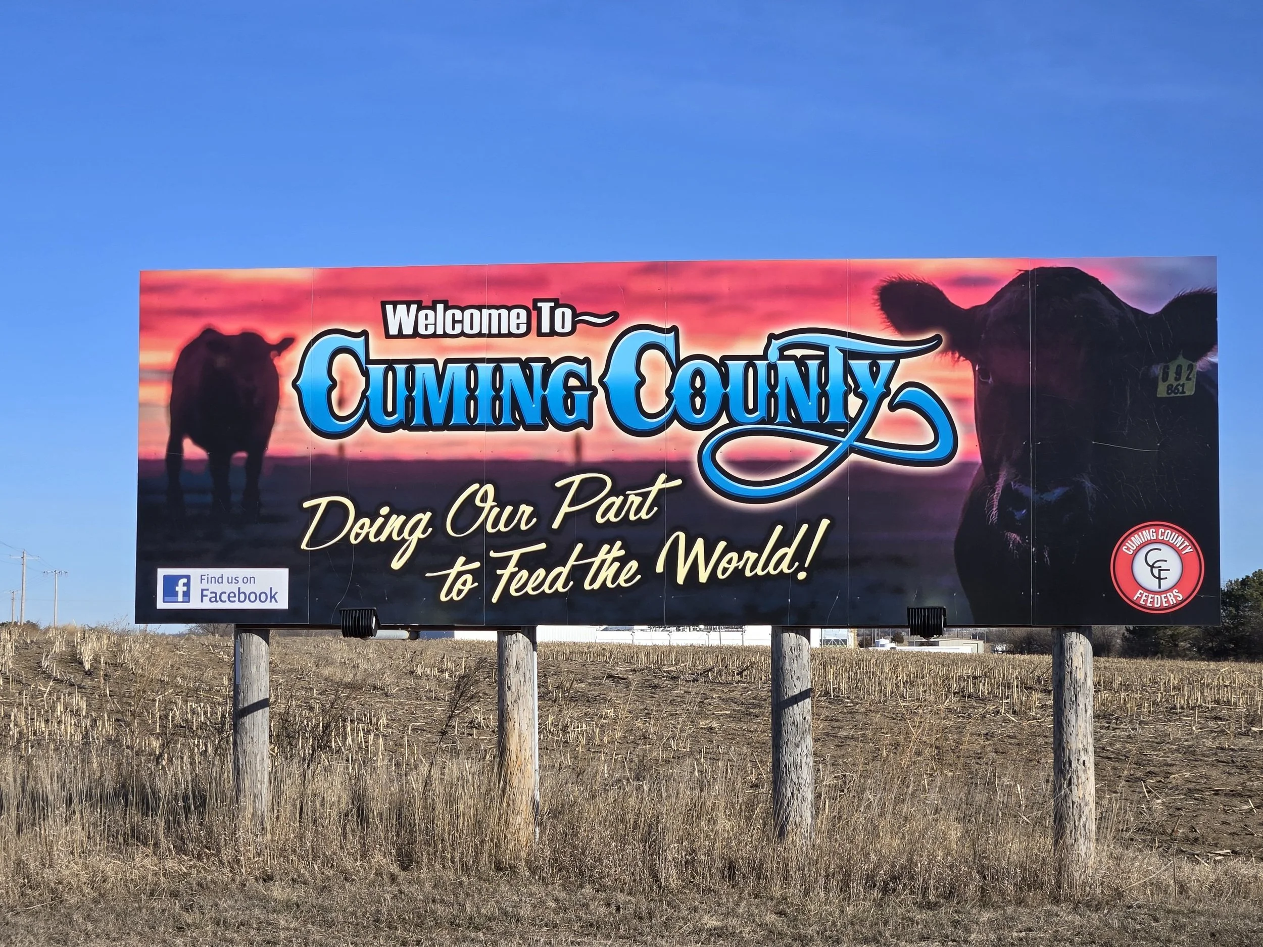 A billboard welcoming visitors to Coming County with a background of a sunset sky and silhouette of a buffalo, and a cow's face on the right side. The billboard also features text promoting feeding the world and a Facebook logo.