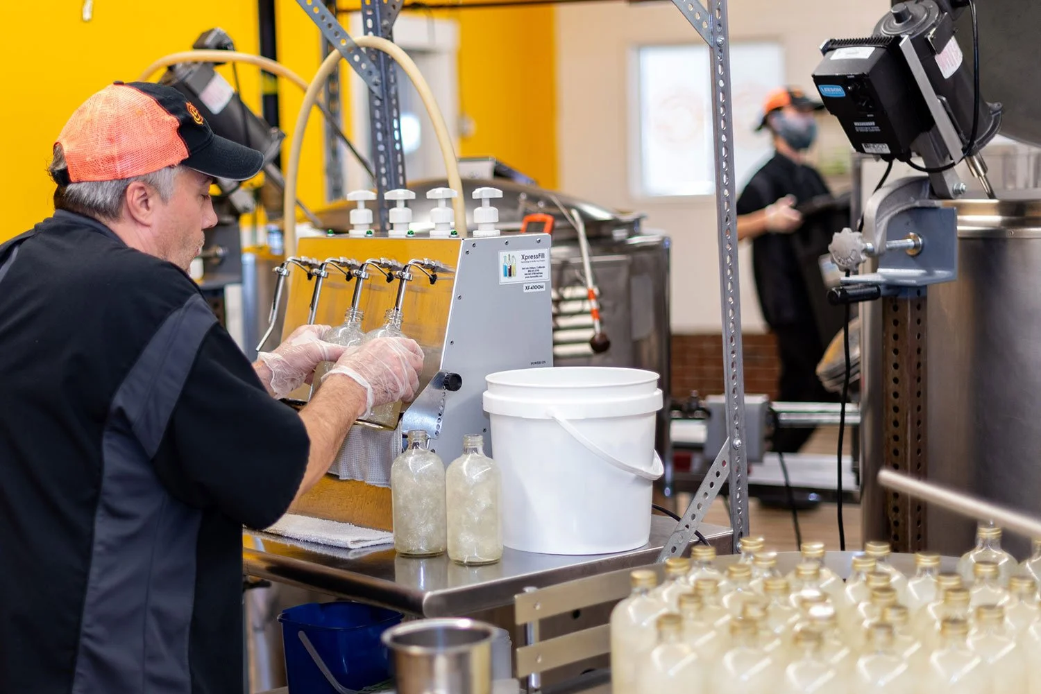A man in a baseball cap and gloves is filling bottles with a liquid from a machine in a brewery or beverage manufacturing setting. Another worker is visible in the background.