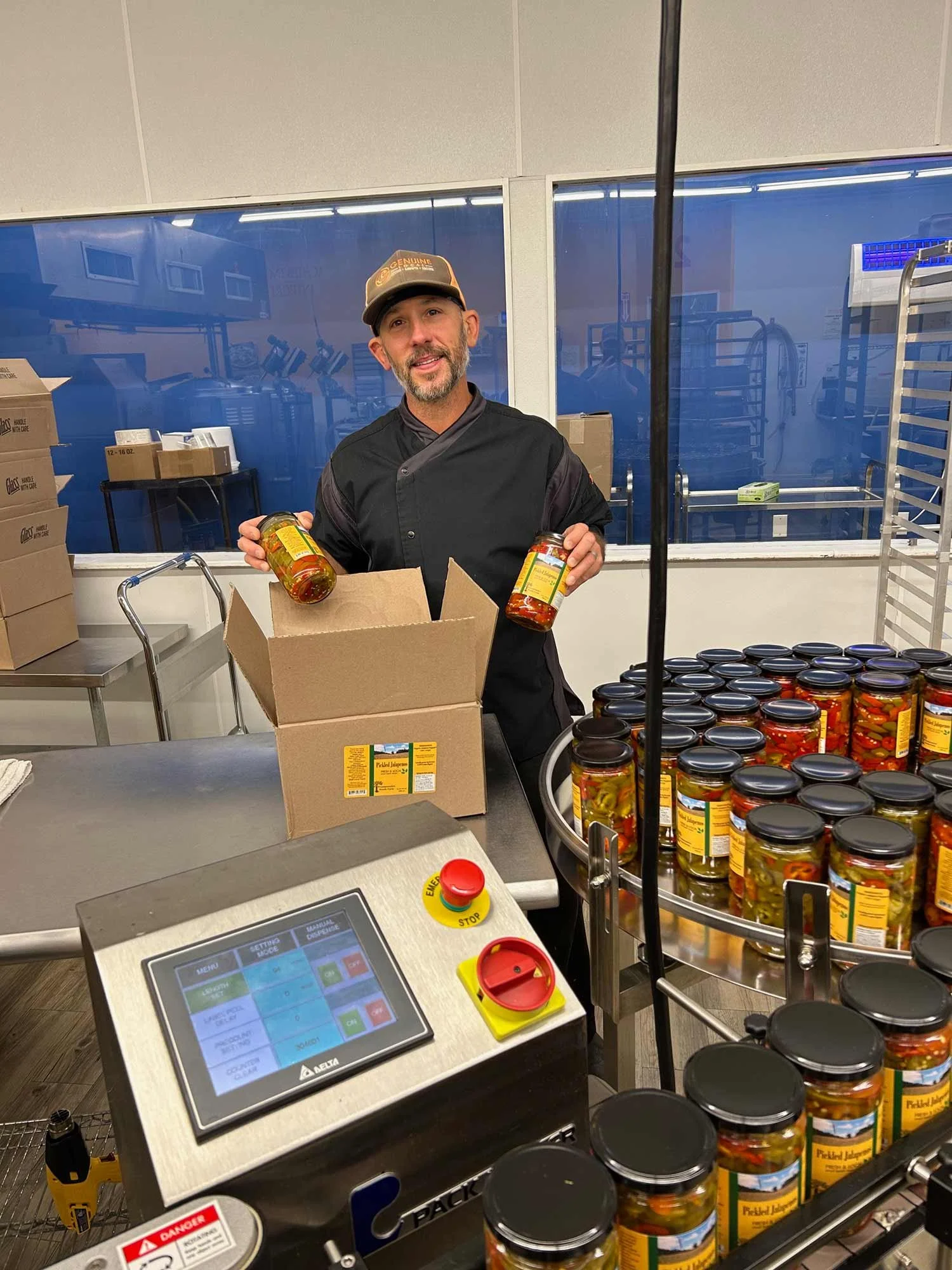 A man in a black uniform and cap standing behind a counter, holding jars of pickled jalapenos, with more jars of pickled jalapenos on a rotating tray, cardboard boxes, and a touchscreen register in front of him.
