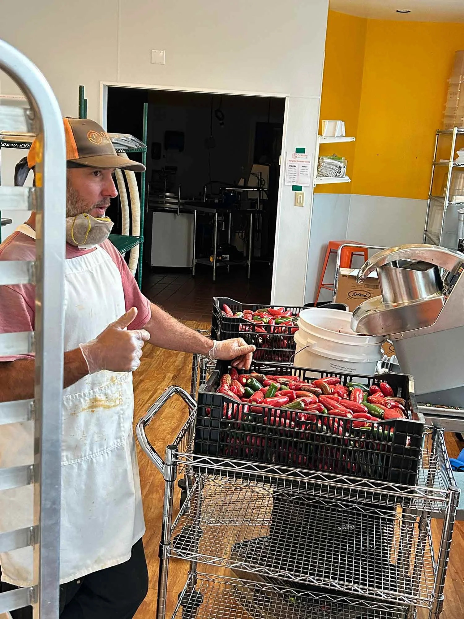 A man in a baseball cap, apron, and gloves standing next to crates of colorful Roma tomatoes in a kitchen or food processing area.