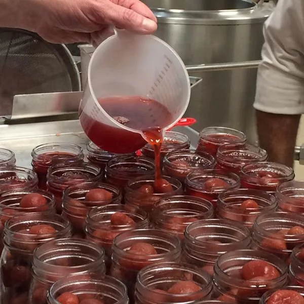 Person pouring red liquid, possibly jam or syrup, into small glass jars filled with round fruits, in a kitchen or processing area.