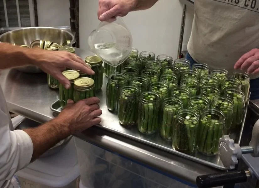 People filling glass jars with sliced cucumbers for pickling in a commercial kitchen.