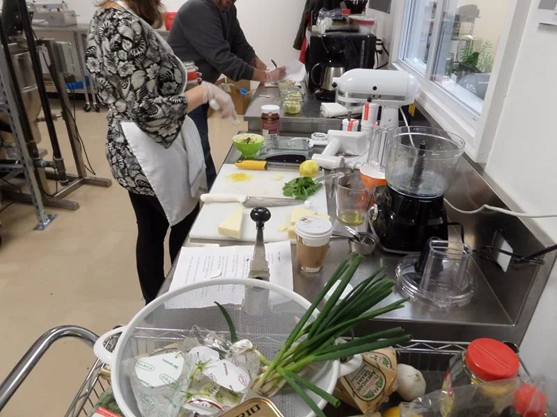 People preparing food in a kitchen or production area with various ingredients and appliances on a stainless steel counter.