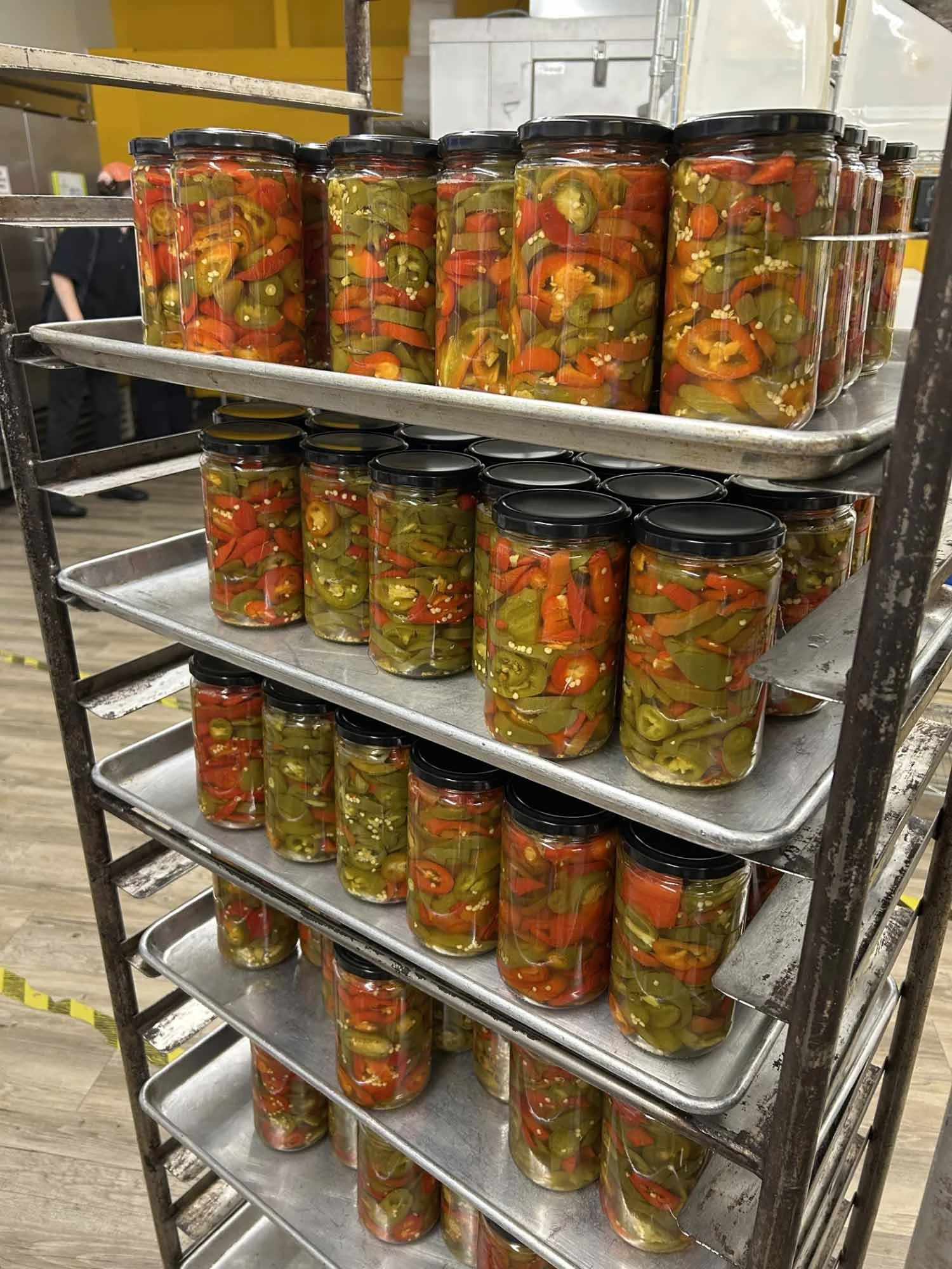 Jars of pickled jalapeño peppers on a metal shelving unit in a commercial kitchen or food processing facility.