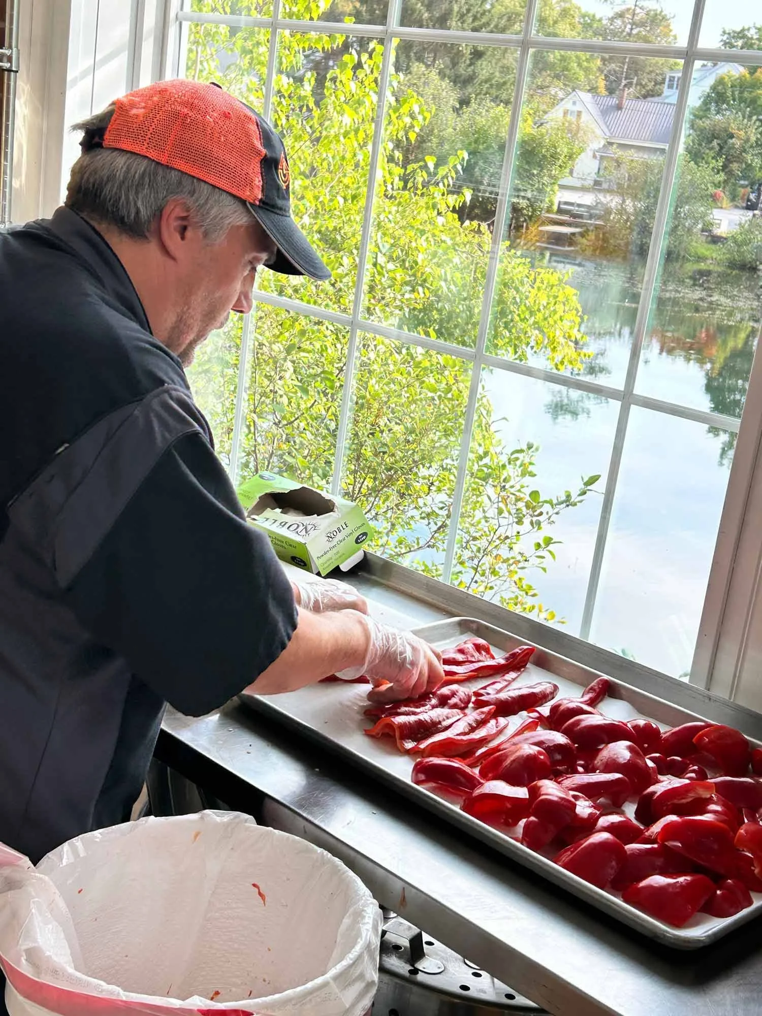 A man wearing a red and black baseball cap, black jacket, and gloves slices red bell peppers on a metal tray by a window overlooking a river and trees.