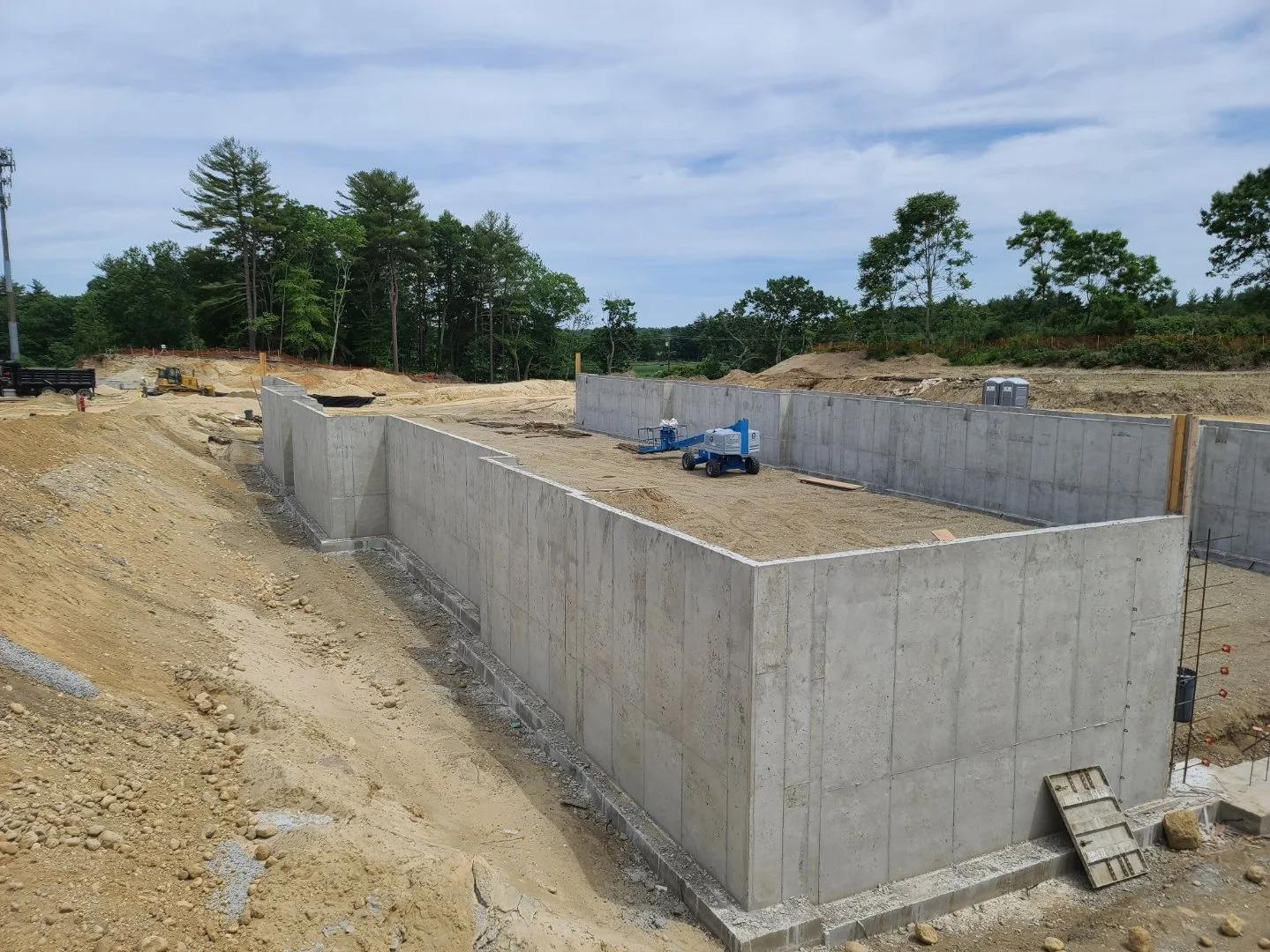 Construction site with concrete foundation walls, construction equipment, and portable toilets, surrounded by dirt with trees in the background.