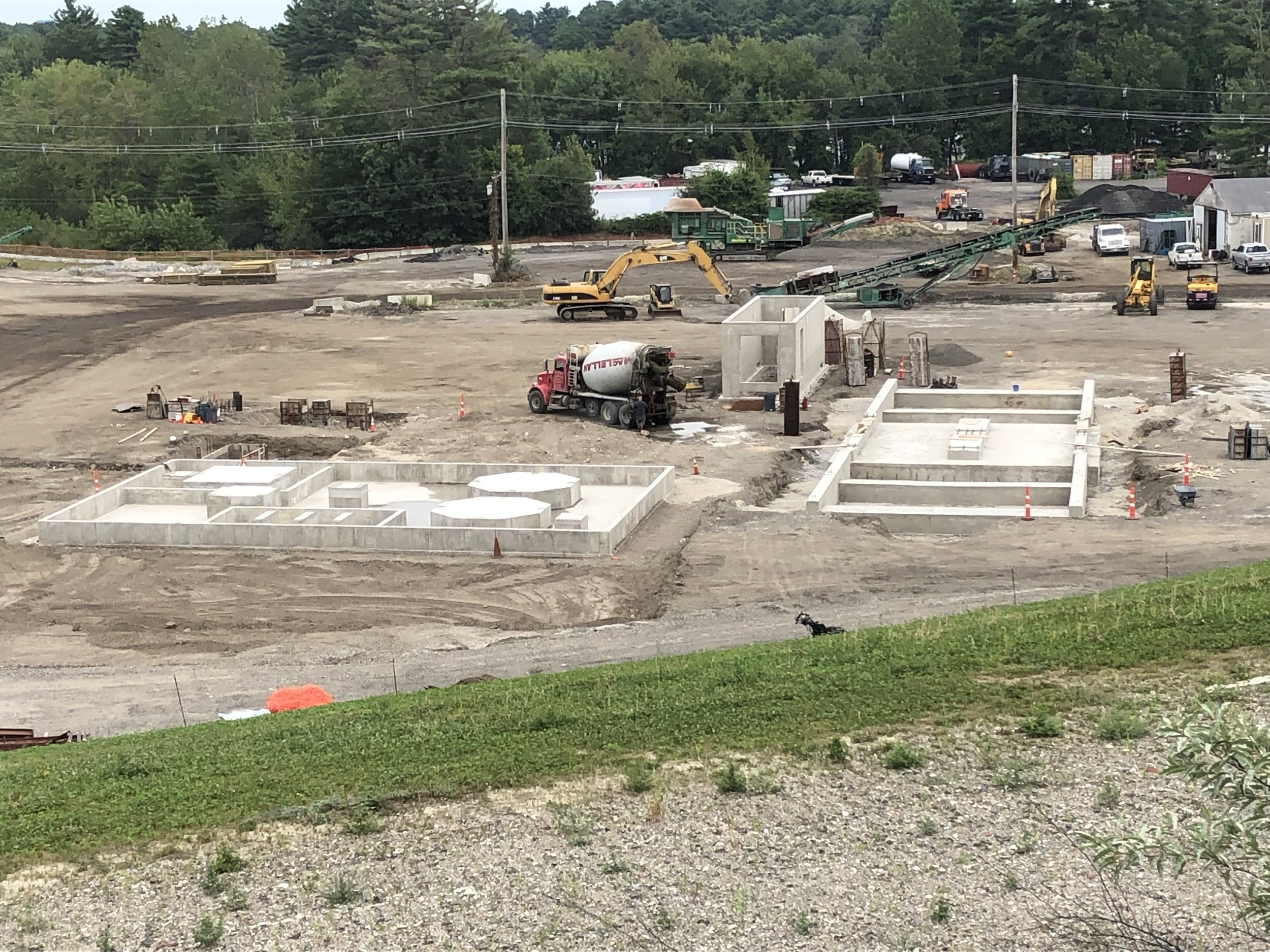 Construction site with concrete foundation structures, construction vehicles, and equipment, with trees and a parking lot in the background.