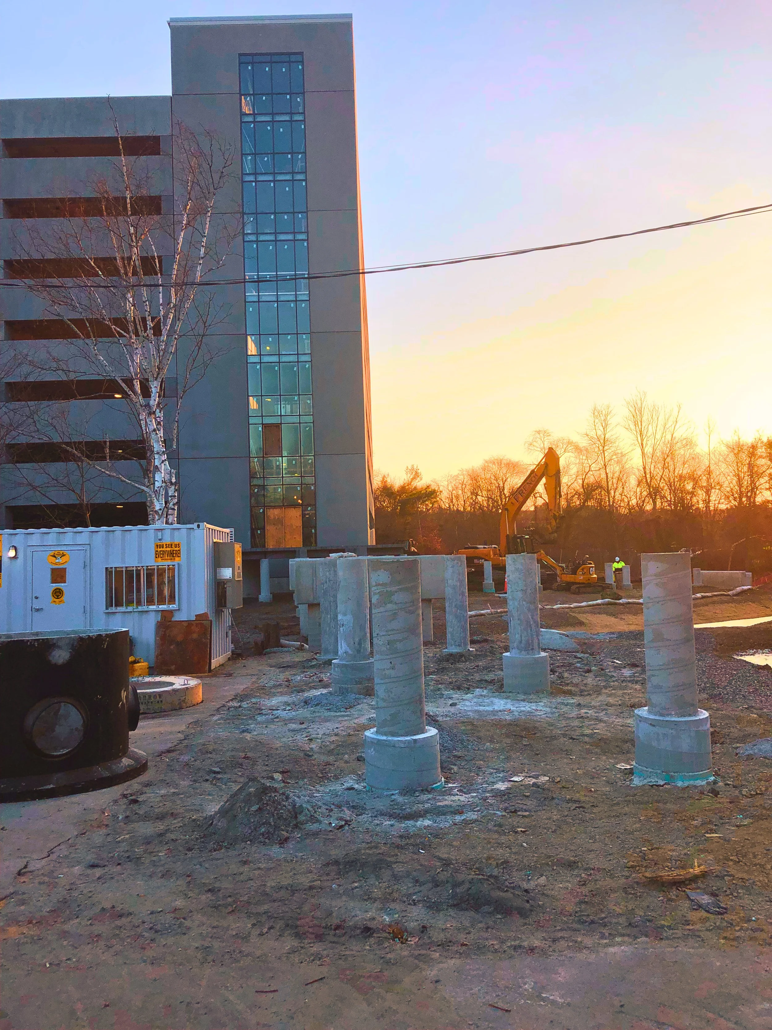 Construction site with concrete pillars, a yellow excavator, and a modern building in the background at sunset.