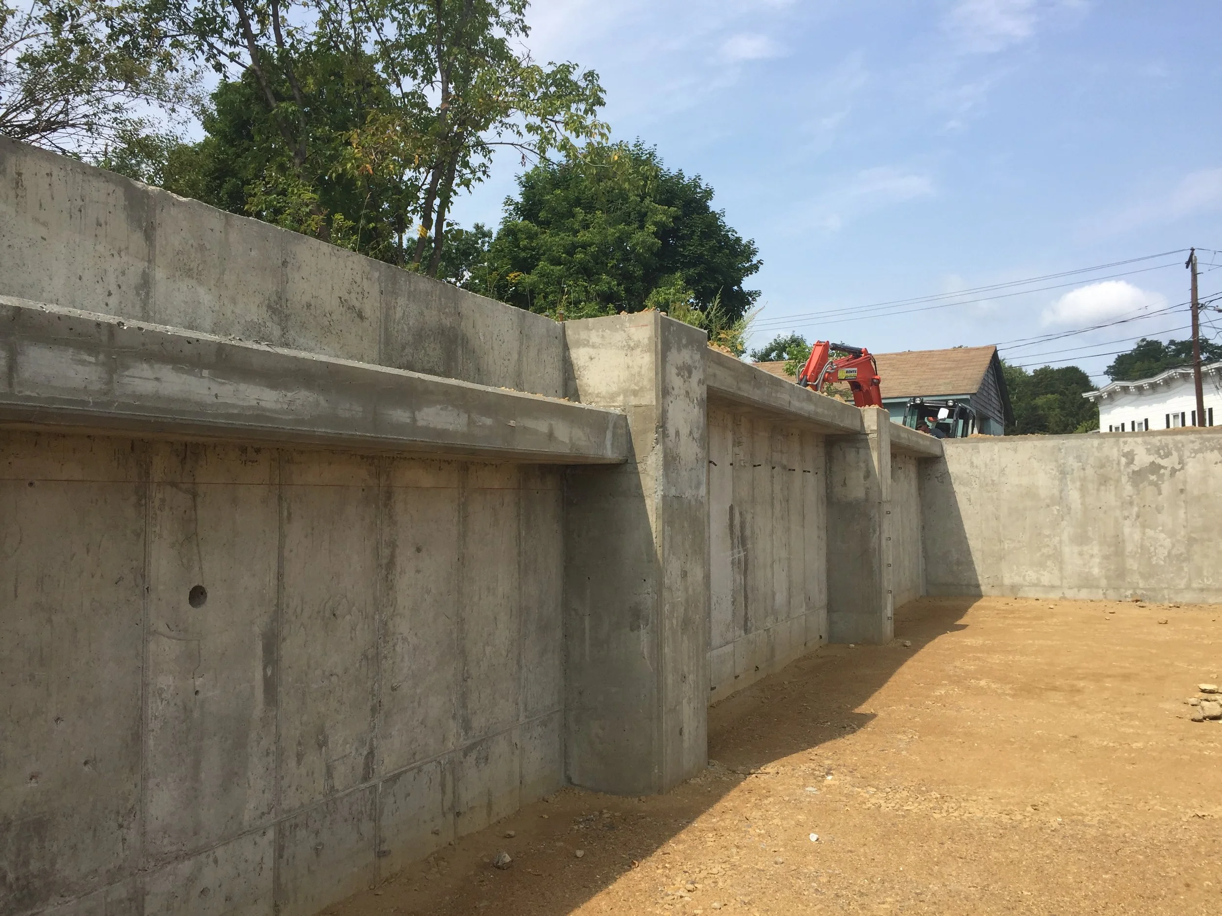 Construction site with concrete walls and a dirt ground, some trees in the background, and construction machinery partially visible.