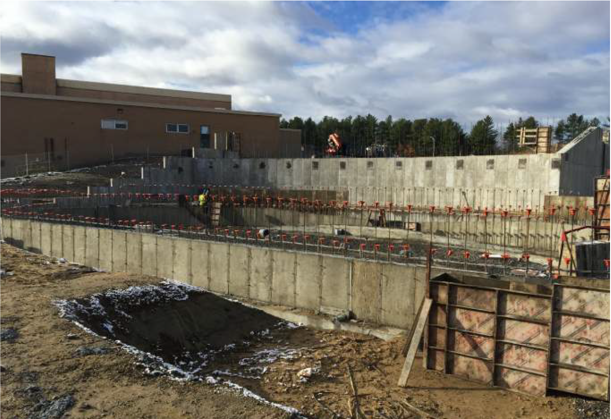Construction site with concrete foundation walls, workers, and construction equipment under cloudy sky.