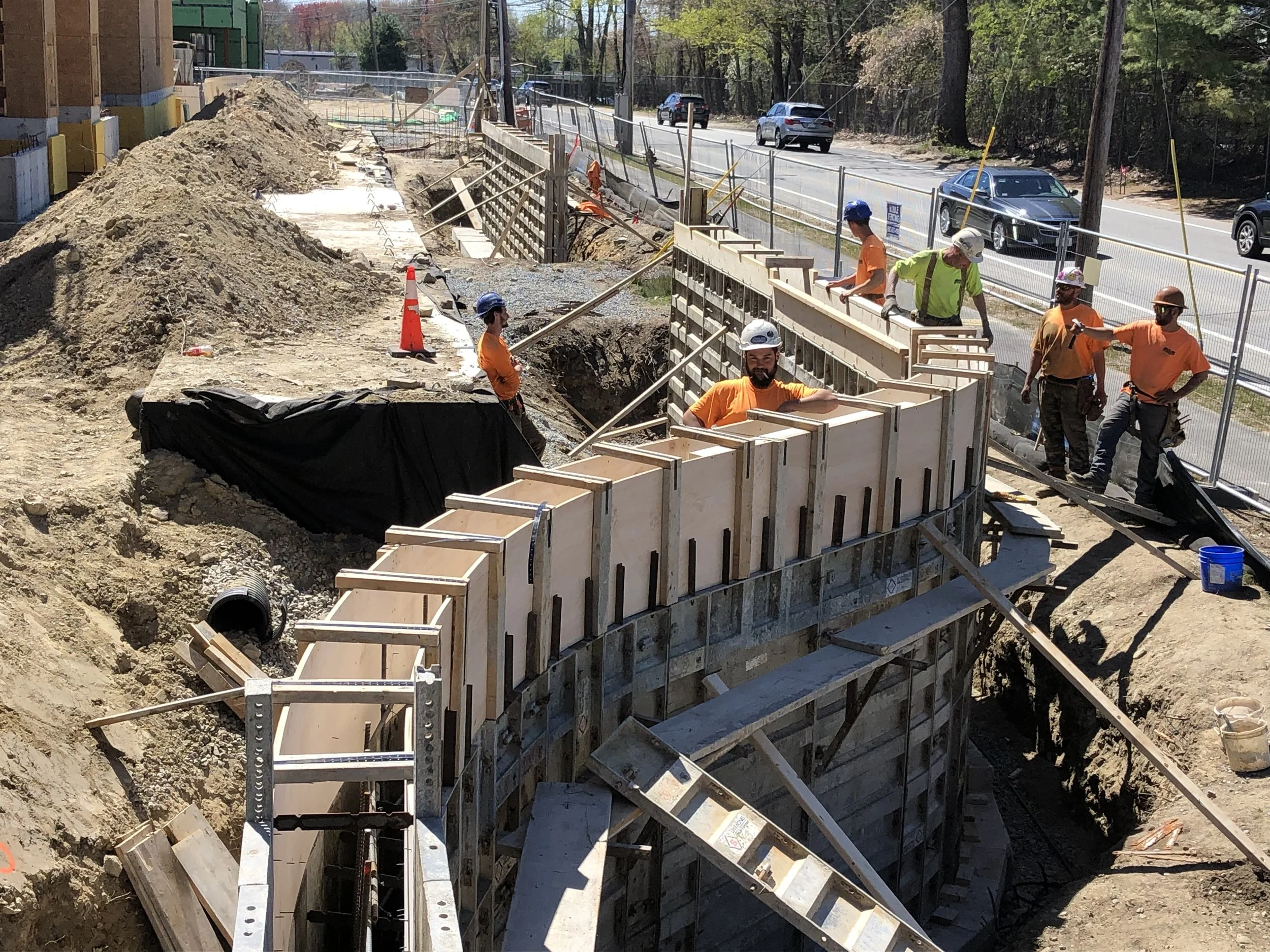 Construction workers in orange shirts and hard hats building a concrete wall on a construction site next to a road, with cars driving by in the background.
