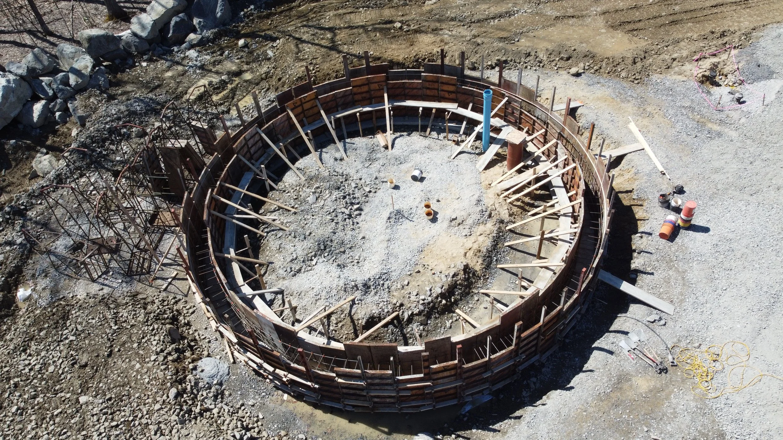 A circular construction site with formwork and scaffolding, surrounded by rocks and dirt, with construction materials scattered around.