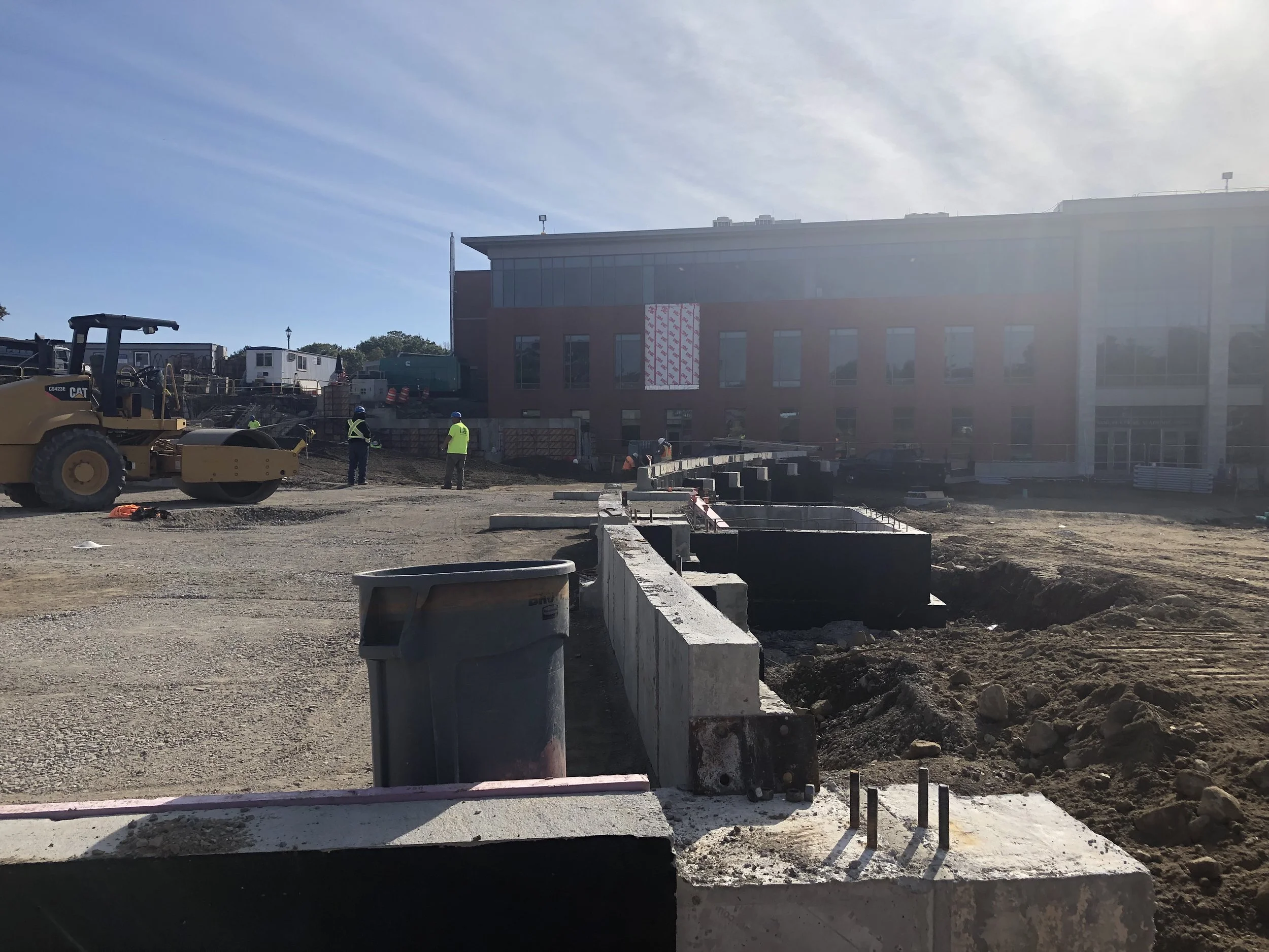 Construction site with workers, heavy machinery, and building structures under development in front of a multi-story red brick building.