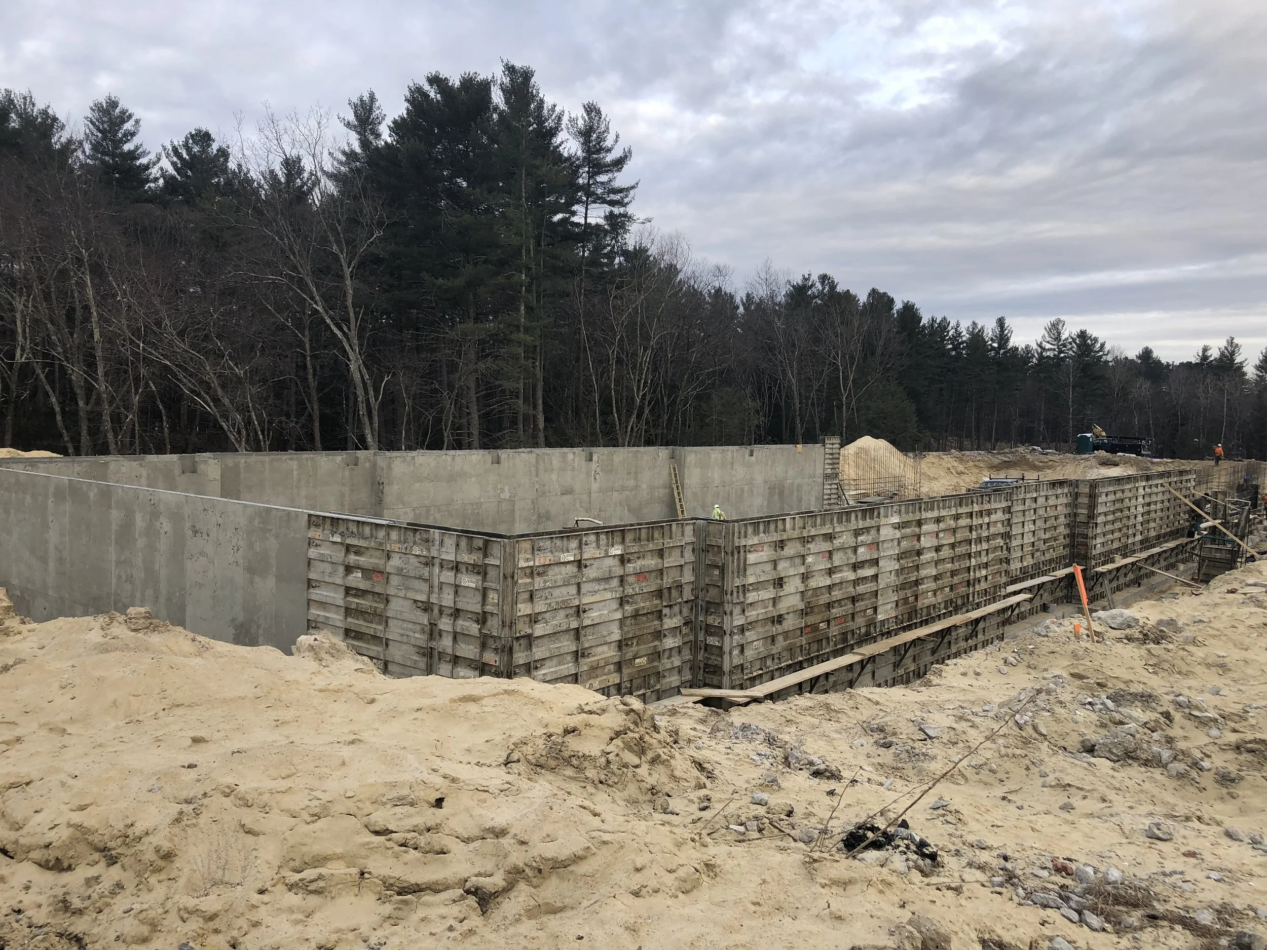 Construction site with concrete wall foundation and wooden formwork, surrounded by dirt and sand, with trees and cloudy sky in the background.