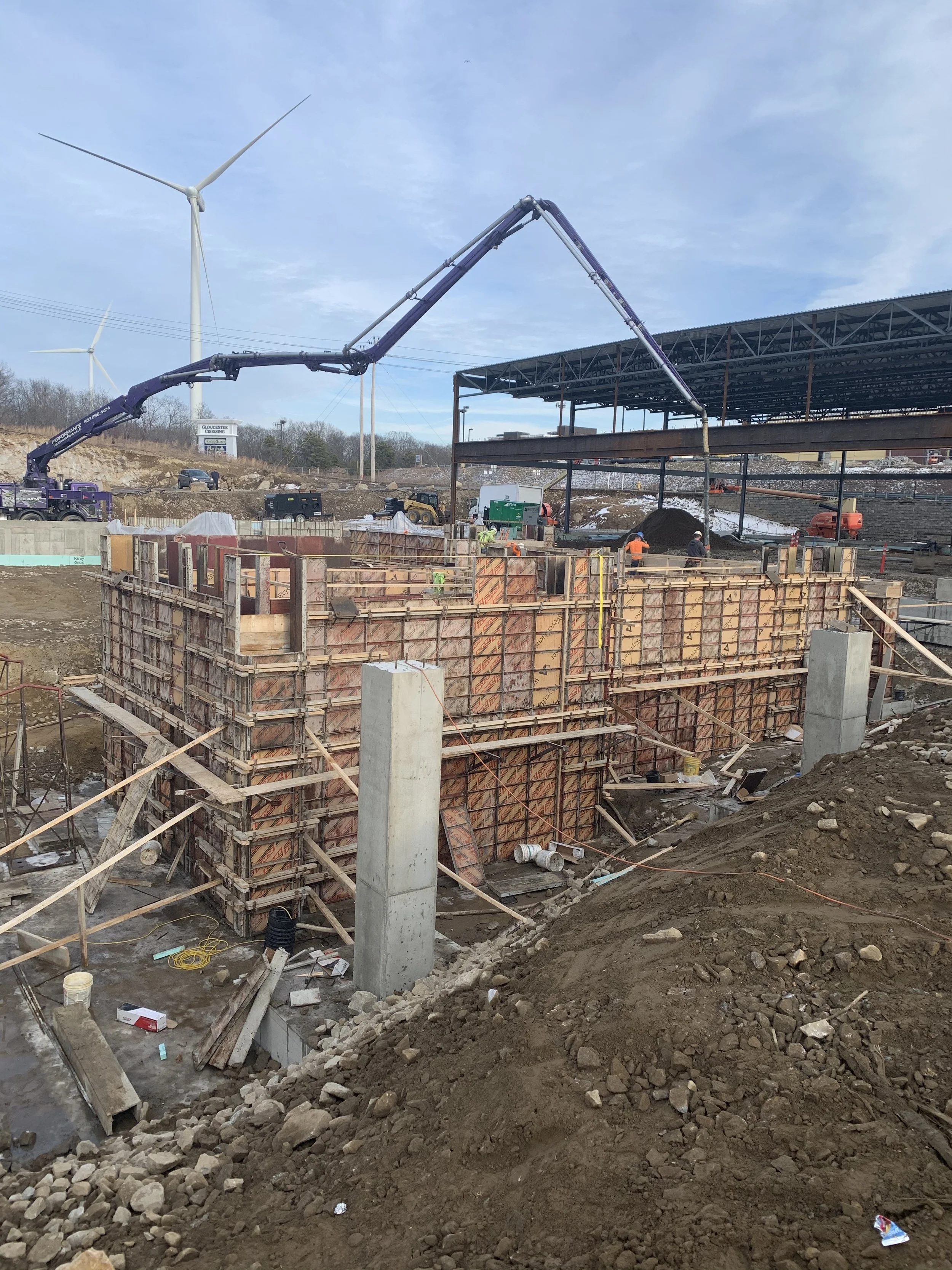Construction site with large wooden formwork, a concrete column, and construction workers, with wind turbines in the background.
