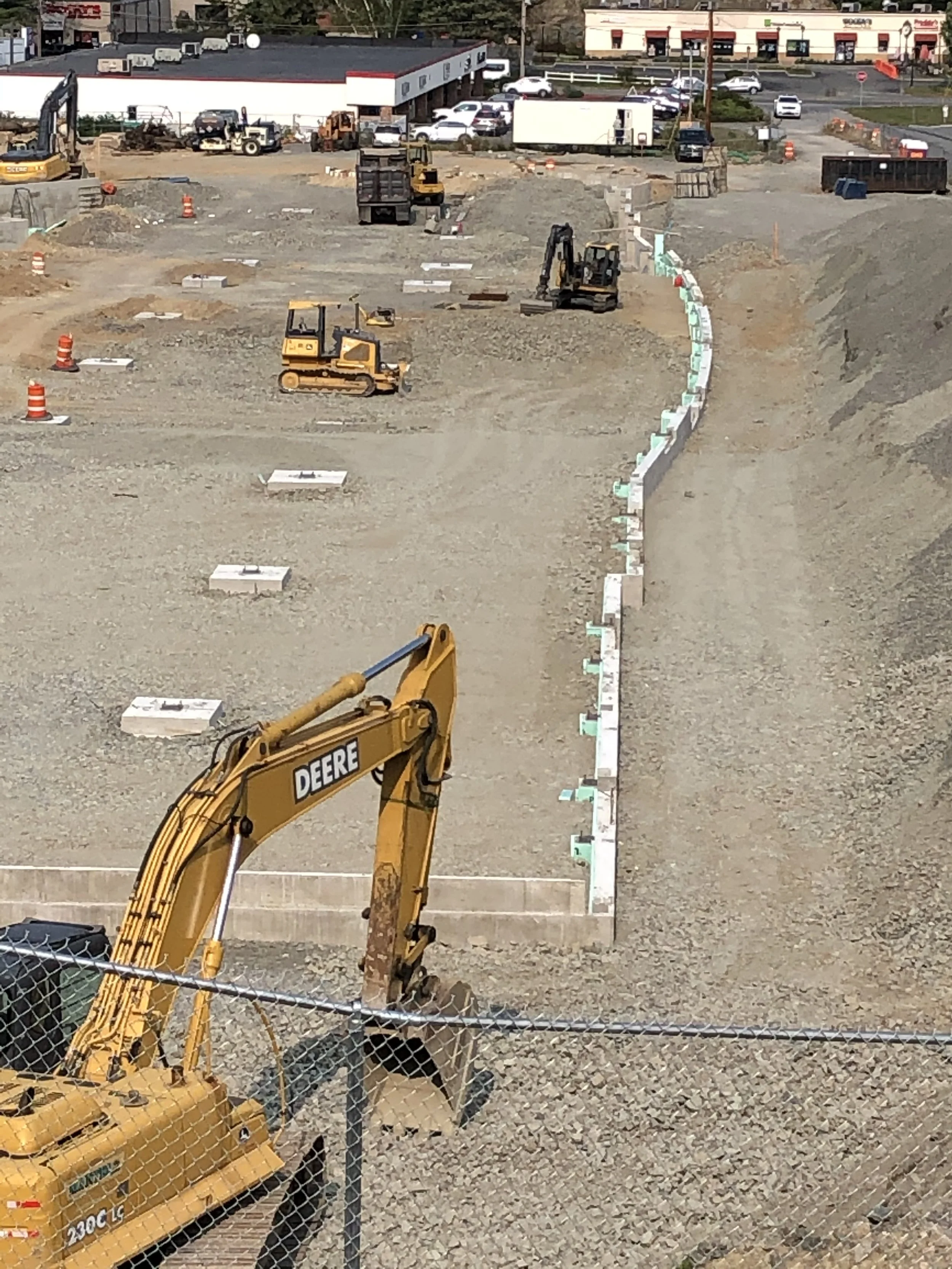 Construction site with heavy machinery, orange traffic cones, and concrete barriers, near a parking lot with cars and a shopping center in the background.