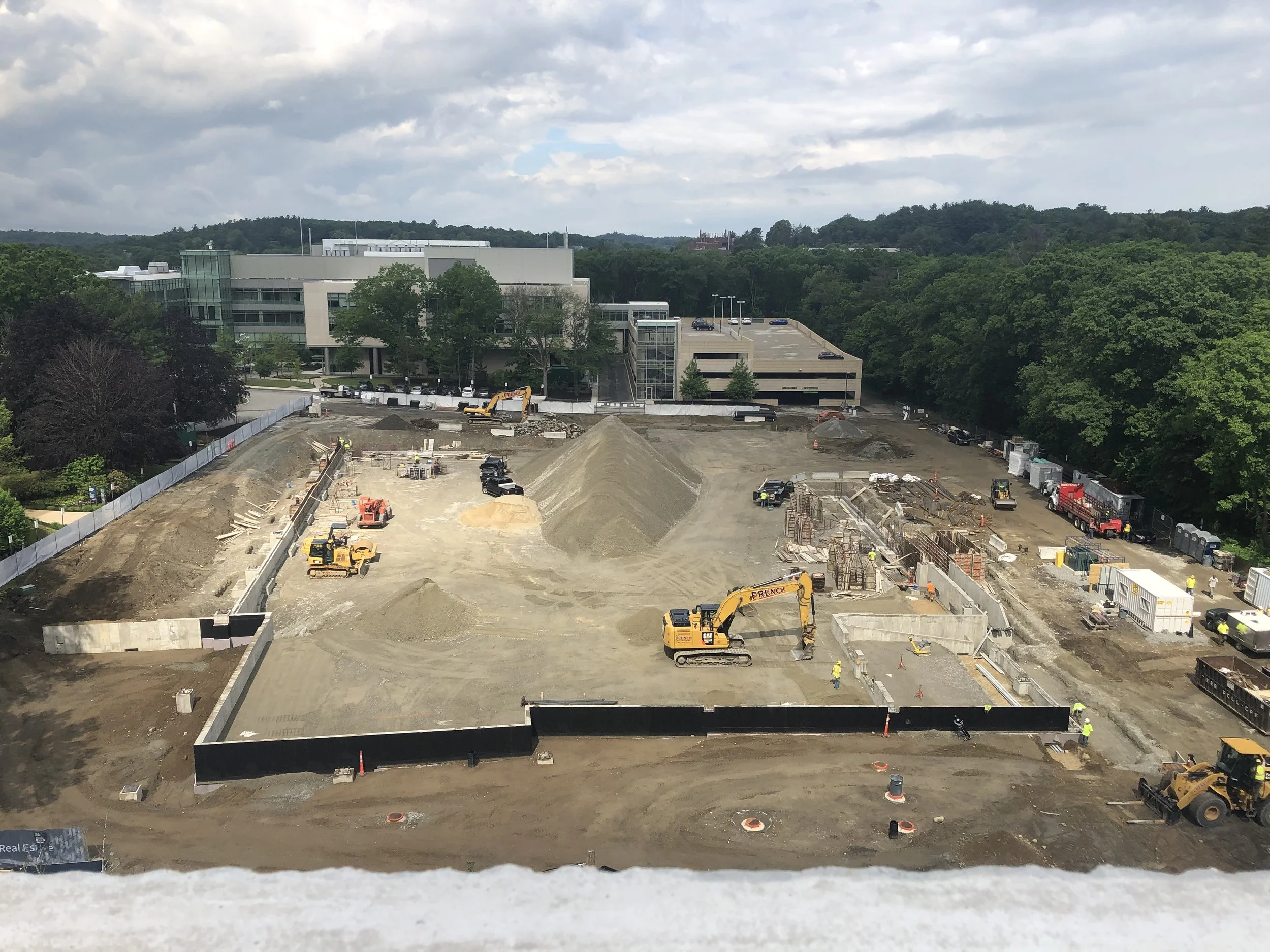 Construction site in progress with heavy machinery and partially built structures, near a modern building with parking lot and trees in the background.