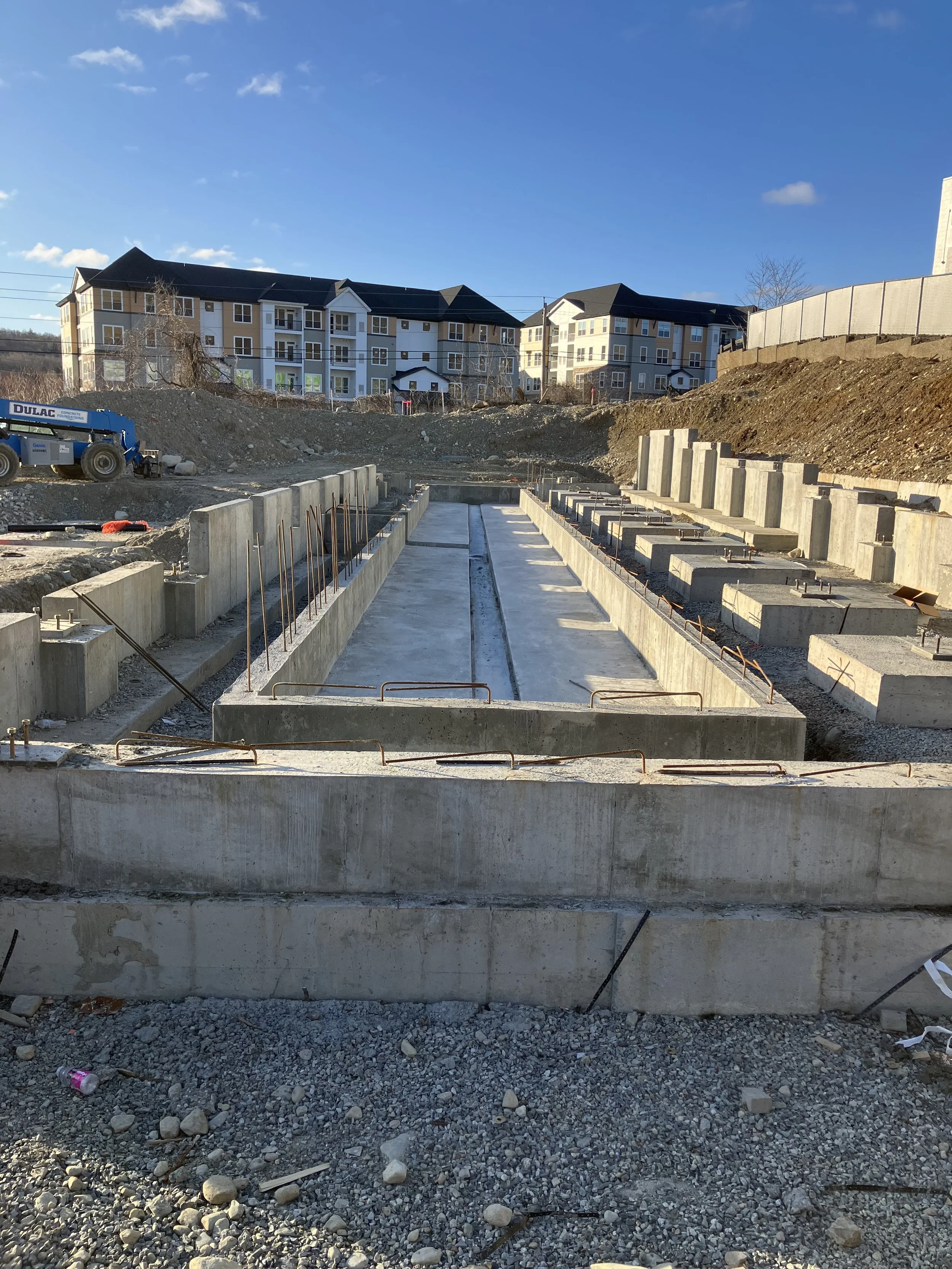 Construction site with concrete foundation and building materials, residential buildings in the background, clear blue sky.