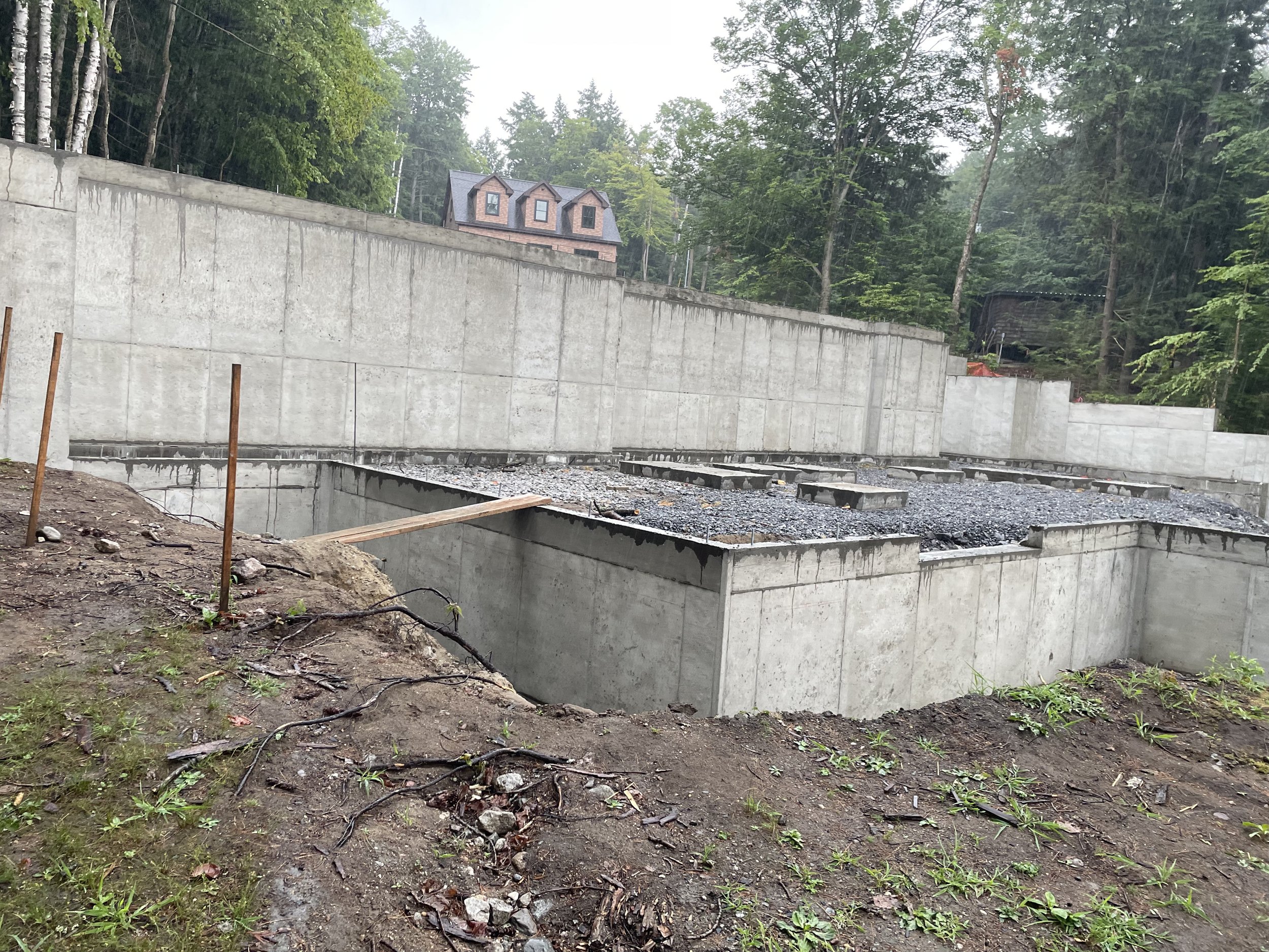 Construction site with concrete foundation and retaining wall, surrounded by trees, with a house and forest in the background.