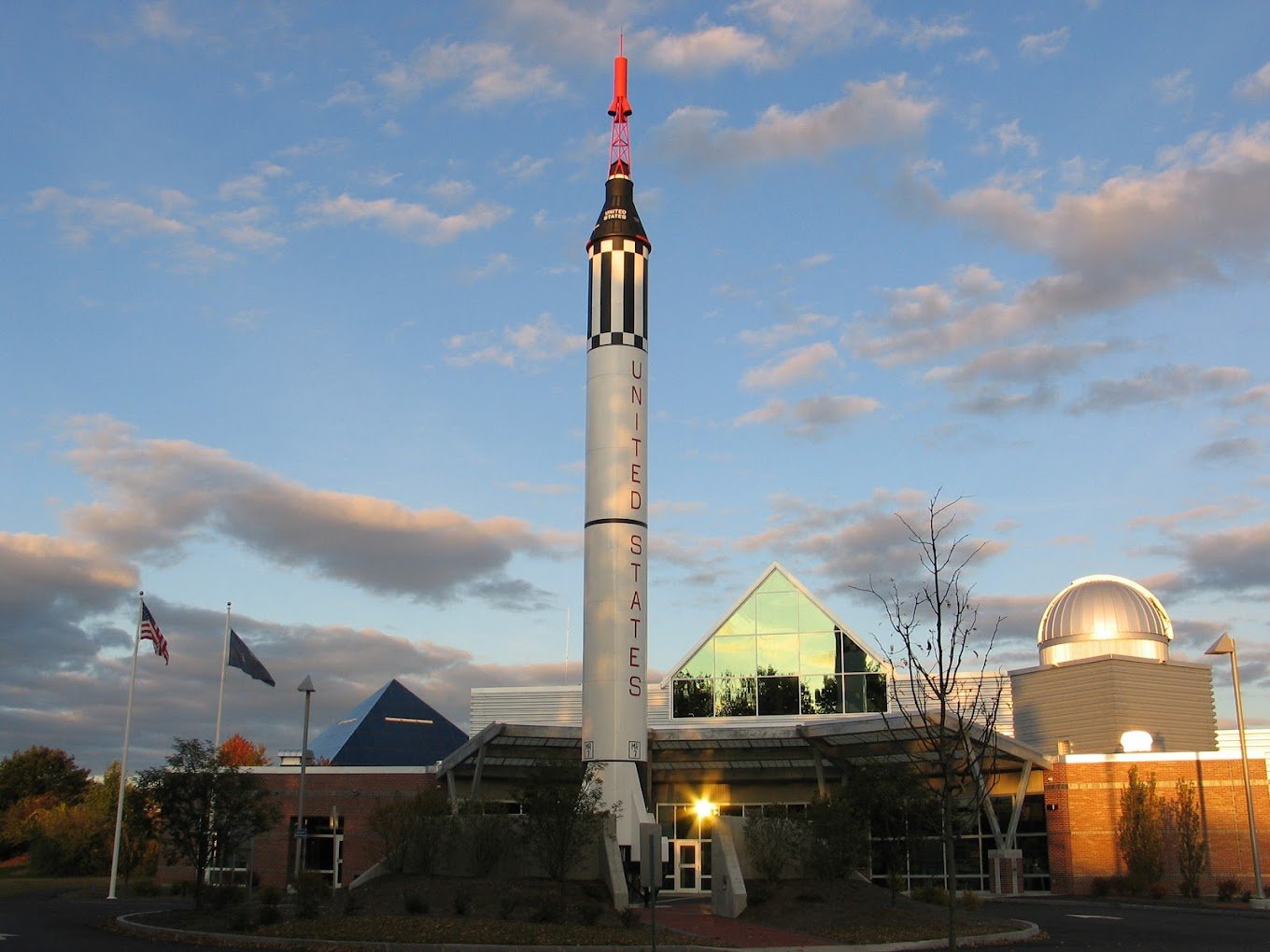 An American space museum with a tall rocket display outside, labeled 'United States,' situated near a modern building with glass windows and a dome-shaped observatory, under a partly cloudy sky.