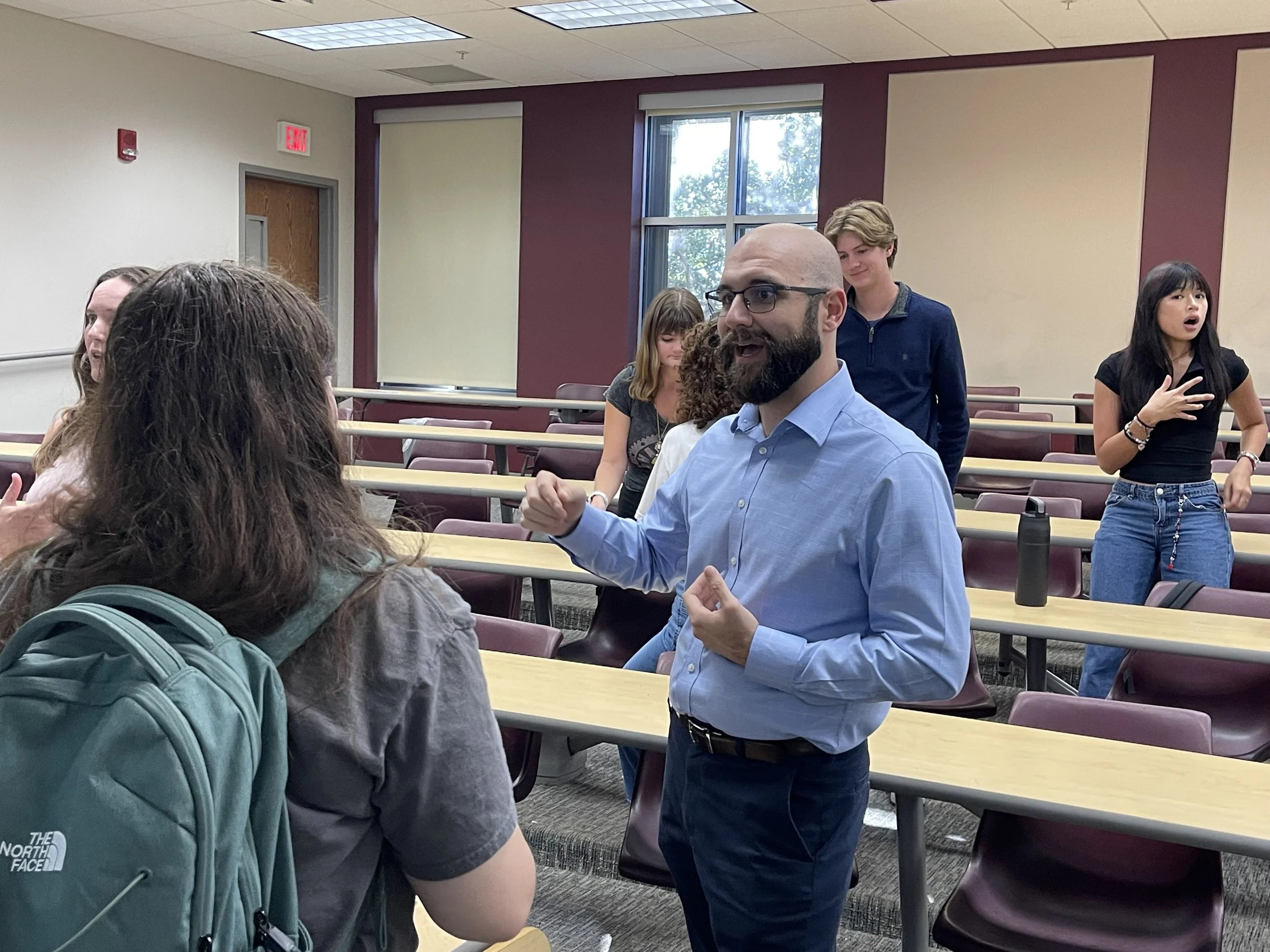 A classroom setting with students and a teacher. The teacher is a bald man with a beard, glasses, dressed in a light blue shirt, engaging in a discussion. Students are seated and standing, some are speaking or listening, with one girl wearing a backp