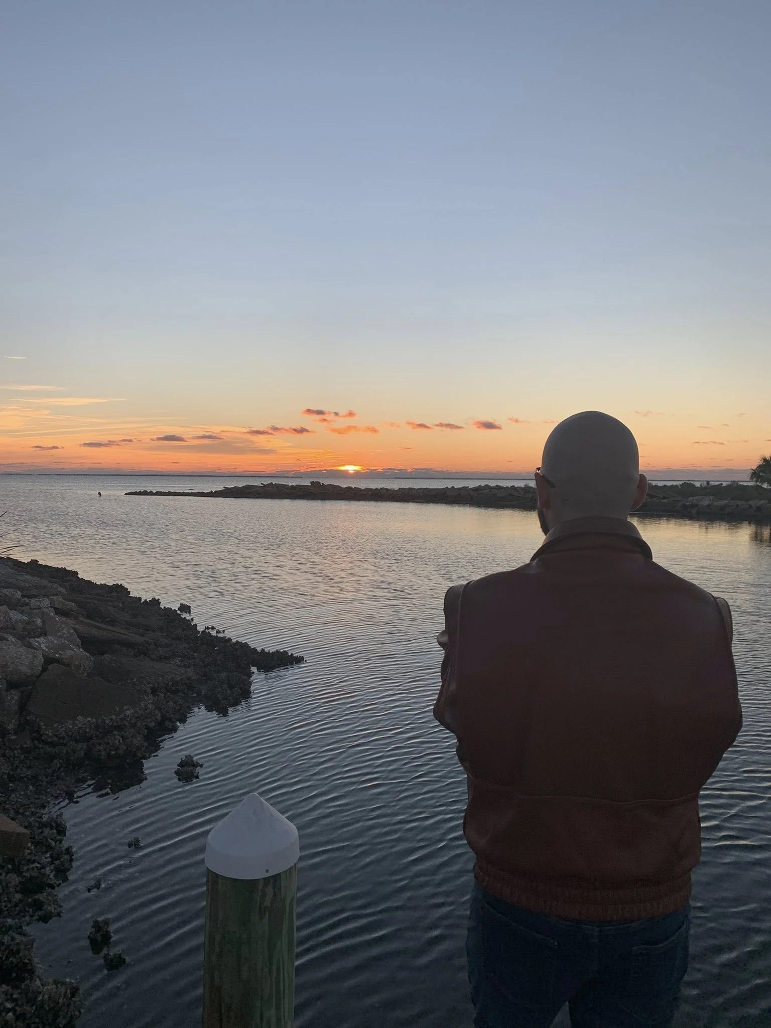 A person with a shaved head wearing a brown jacket stands near a body of water, watching a sunset with orange and pink hues on the horizon.