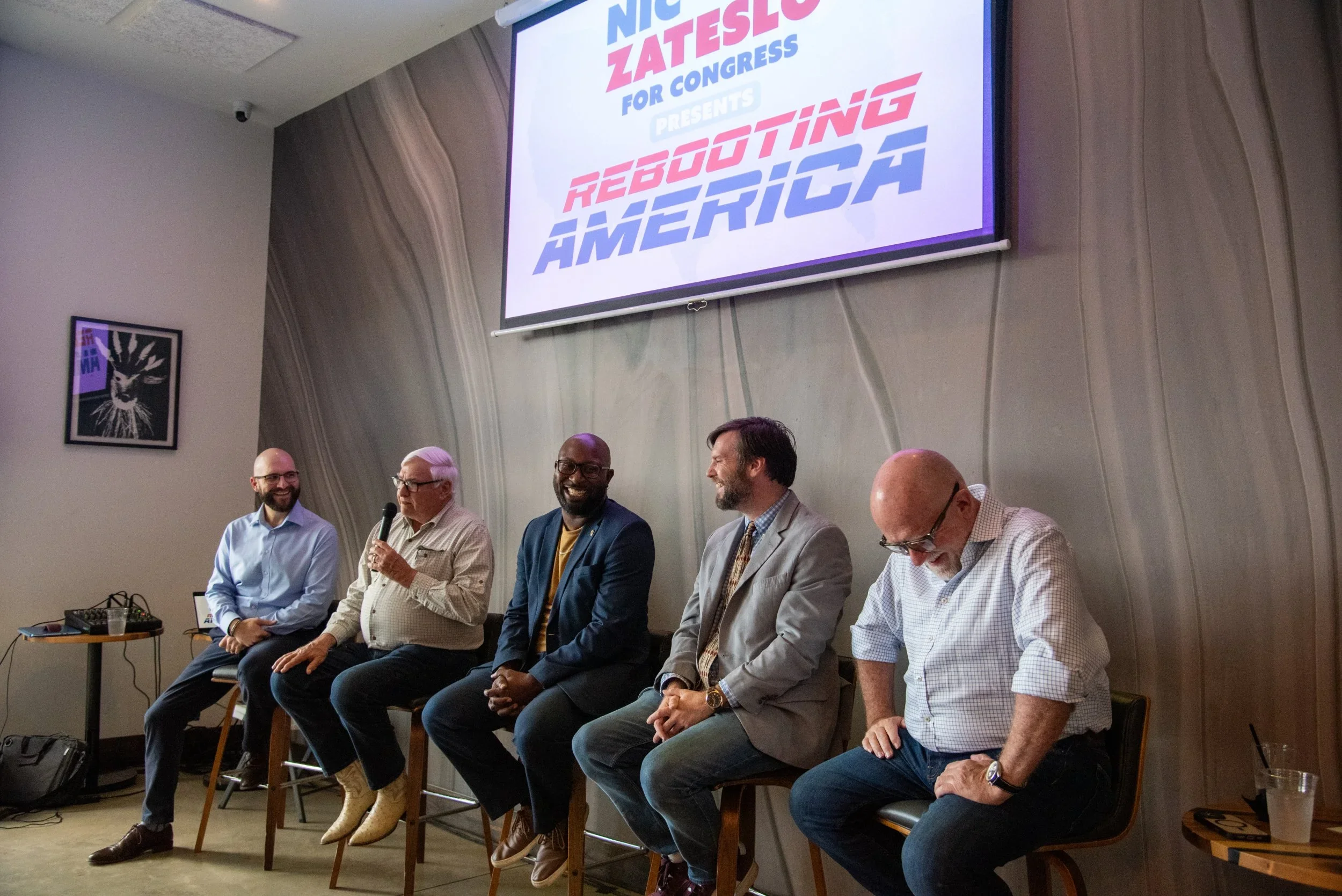 Six men sitting on chairs at a panel discussion, with a large screen behind them displaying the text 'NIC ZATESS FOR CONGRESS PRESENTS REBOOTING AMERICA'. One man holds a microphone, and there is a framed picture on the wall to the left.
