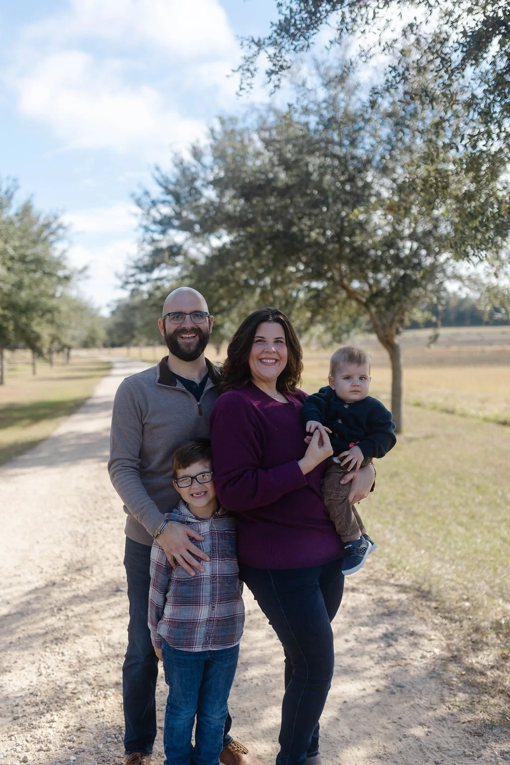 A family of four standing outdoors on a gravel path in a park with trees and open field in the background, smiling and posing for the photo.