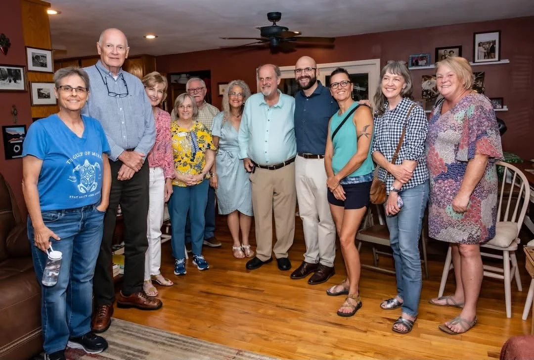 Group of twelve adults gathered together indoors, smiling for a photo, with some holding items like a water bottle and glasses, in a room with wooden floors and decorated walls.